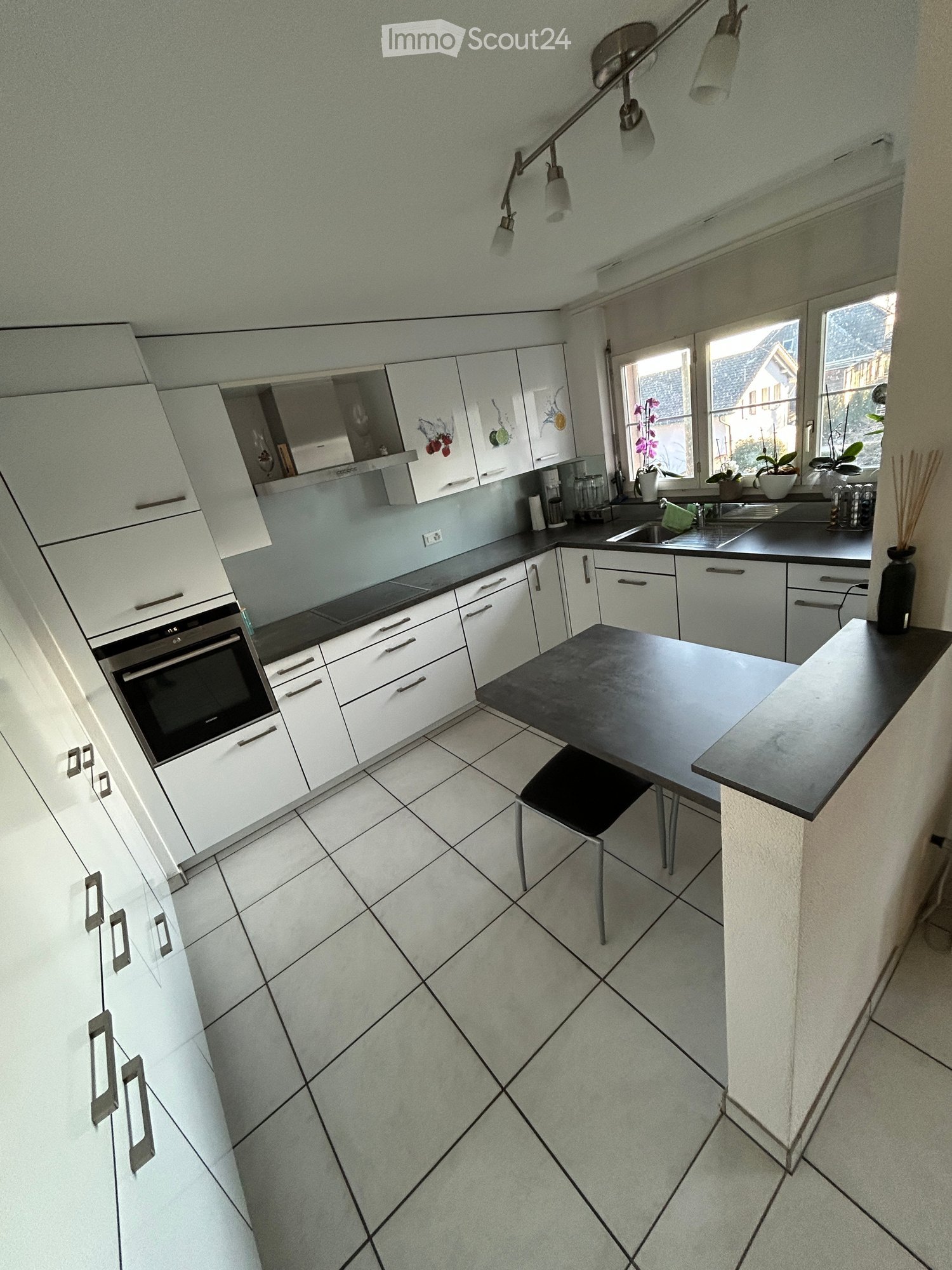 Modern kitchen with white cabinets, black countertops, and a central island with a black table and stool. Features include a sink, oven, and various kitchen appliances.