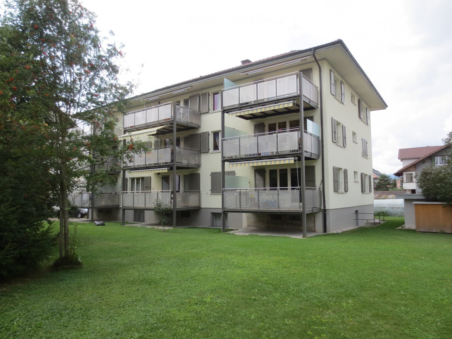 A three-story residential building with multiple balconies, surrounded by a well-maintained lawn and trees.