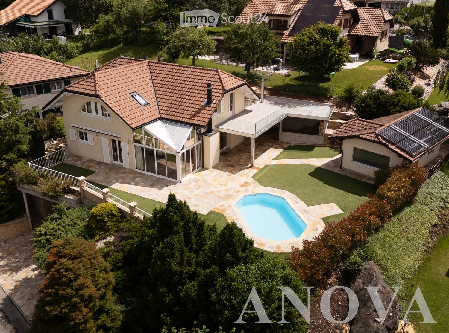 Aerial view of a residential area with multiple houses, a swimming pool, well-maintained gardens, and a patio area with a covered structure.
