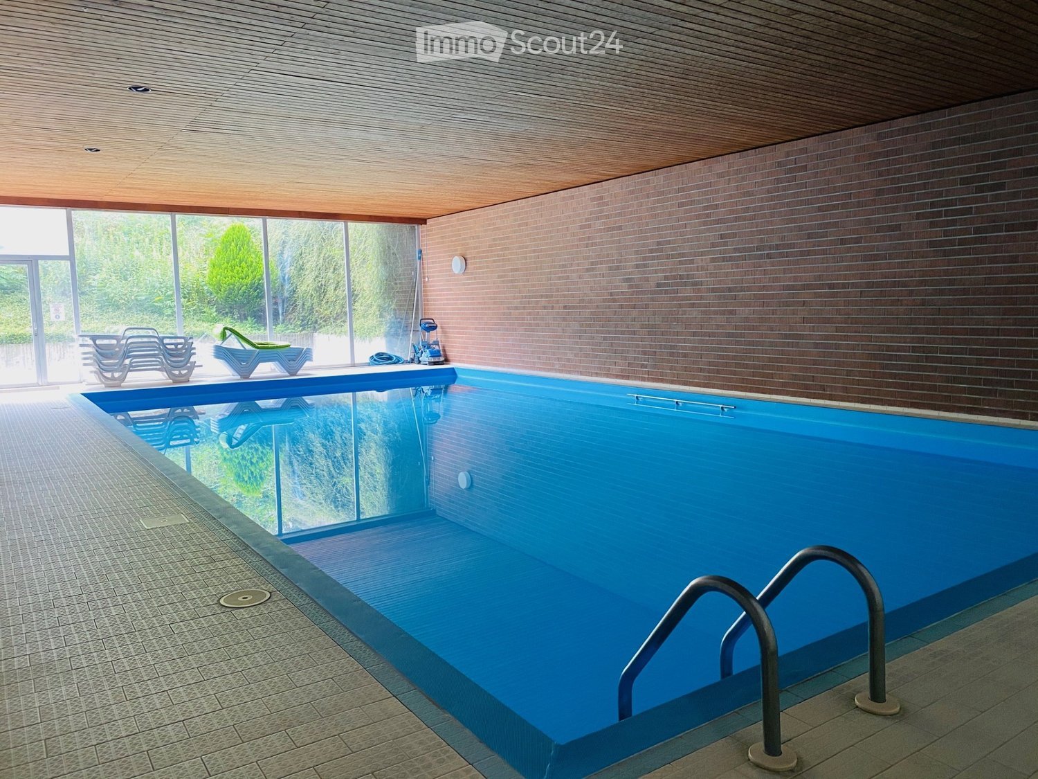 Indoor swimming pool area with clear blue water, surrounded by tiled flooring, wooden ceiling, and large windows offering a view of greenery outside. Lounge chairs are positioned near the pool.