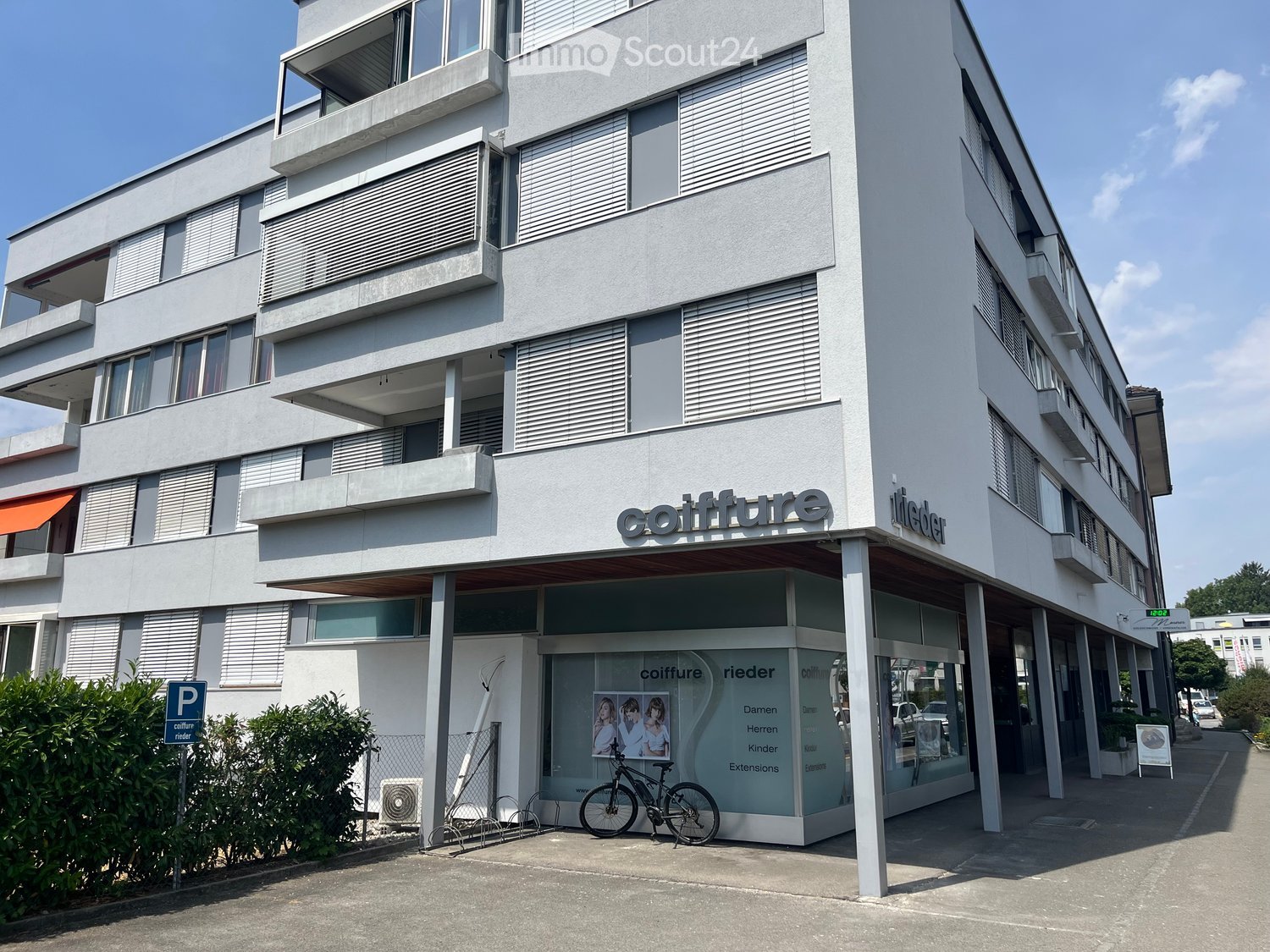 Modern multi-story building with white facade, balconies, and large windows. A storefront with glass windows displays signage for 'coiffure rieder'. A bicycle is parked near the entrance, and a parking sign is visible.