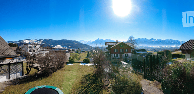 A panoramic view of a residential area with houses, green lawns, trees, and distant snow-capped mountains under a bright blue sky.