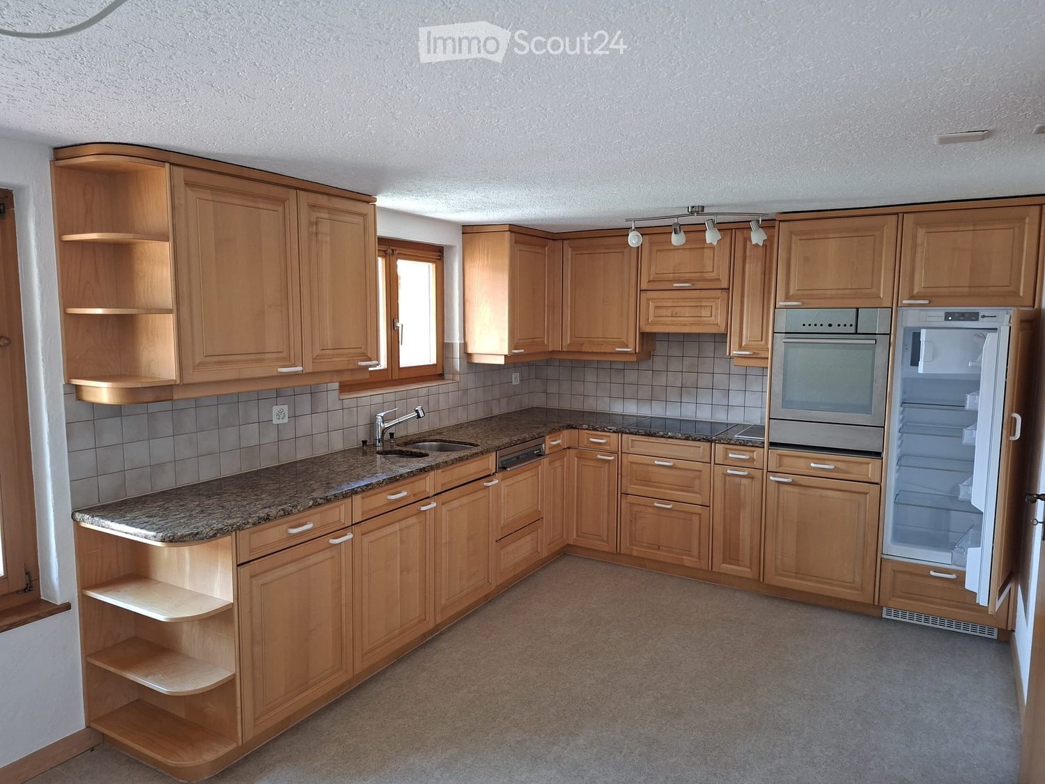 A modern kitchen with light wooden cabinets, granite countertops, and built-in appliances including a sink, oven, and refrigerator.