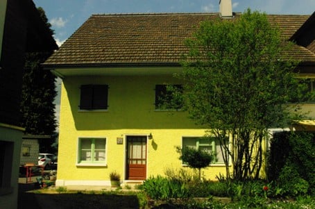 Single-story yellow house with a brown tiled roof, white-framed windows, and a small front garden with trees and shrubs.