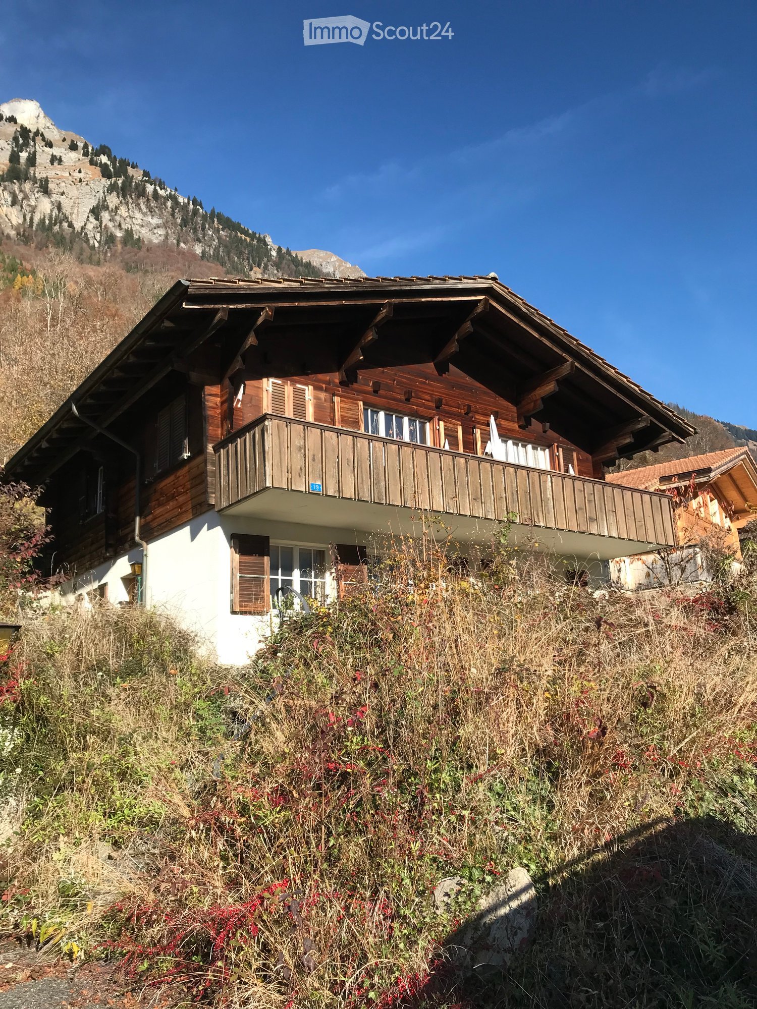 A two-story wooden house with a balcony, situated on a hillside with a mountain view in the background.
