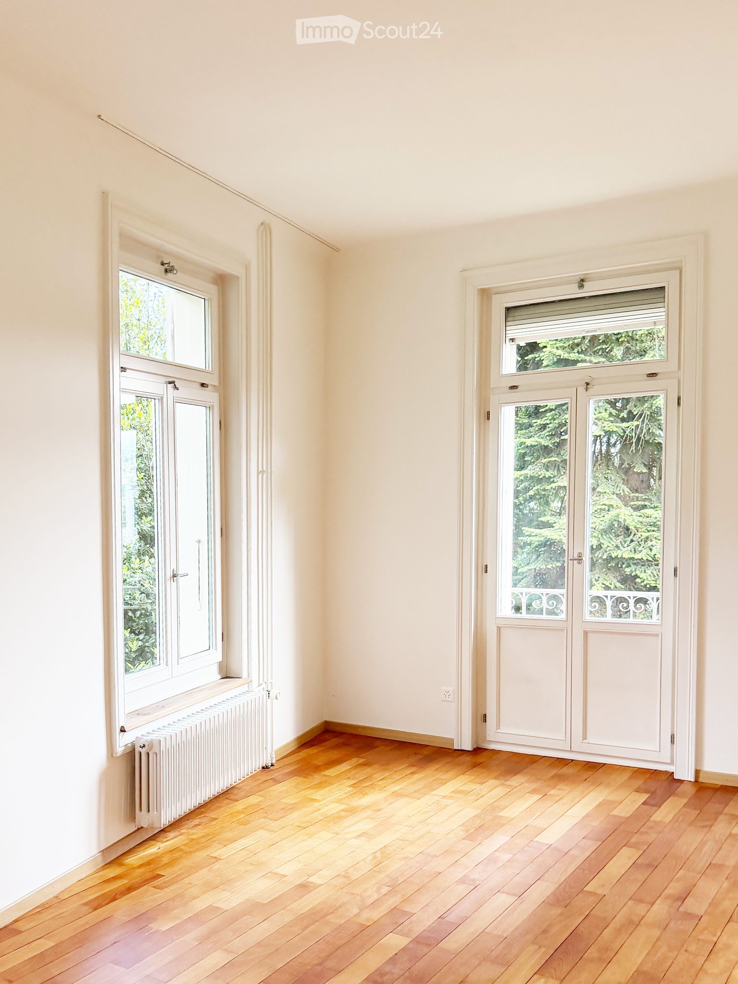 Empty room with light-colored walls, wooden flooring, and large windows with white frames. A radiator is visible under one window, and a set of double doors with glass panels is on the right side.