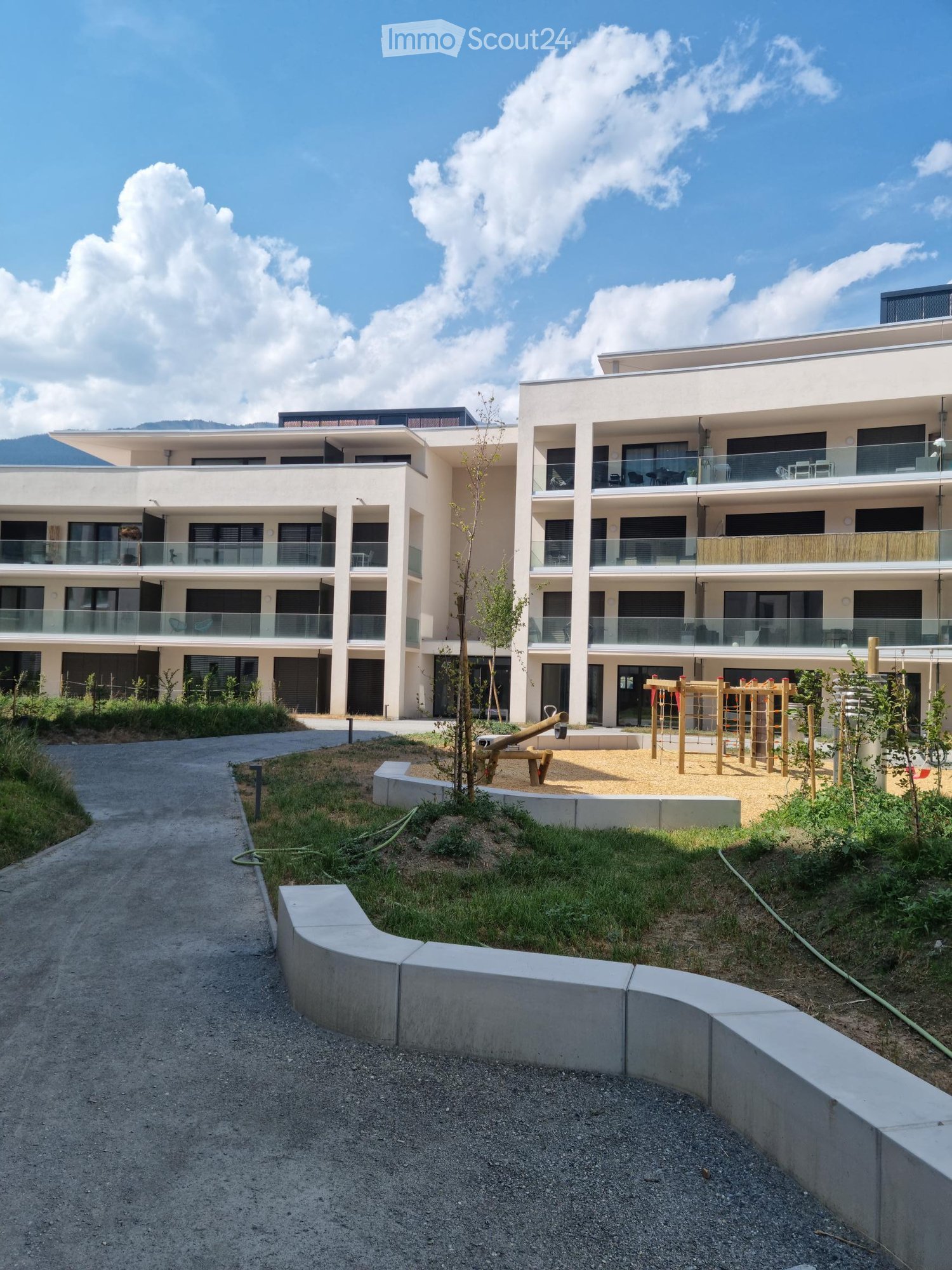 Modern multi-story residential building with balconies, surrounded by a landscaped area with playground equipment and pathways.