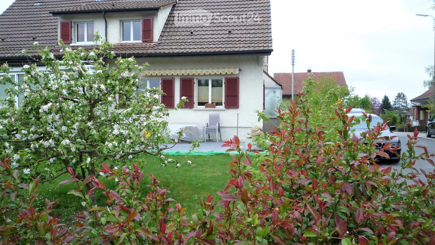 Two-story house with red shutters, white walls, and a tiled roof. The front yard has a well-maintained lawn, flowering trees, and shrubs. A car is parked on the street in front of the house.
