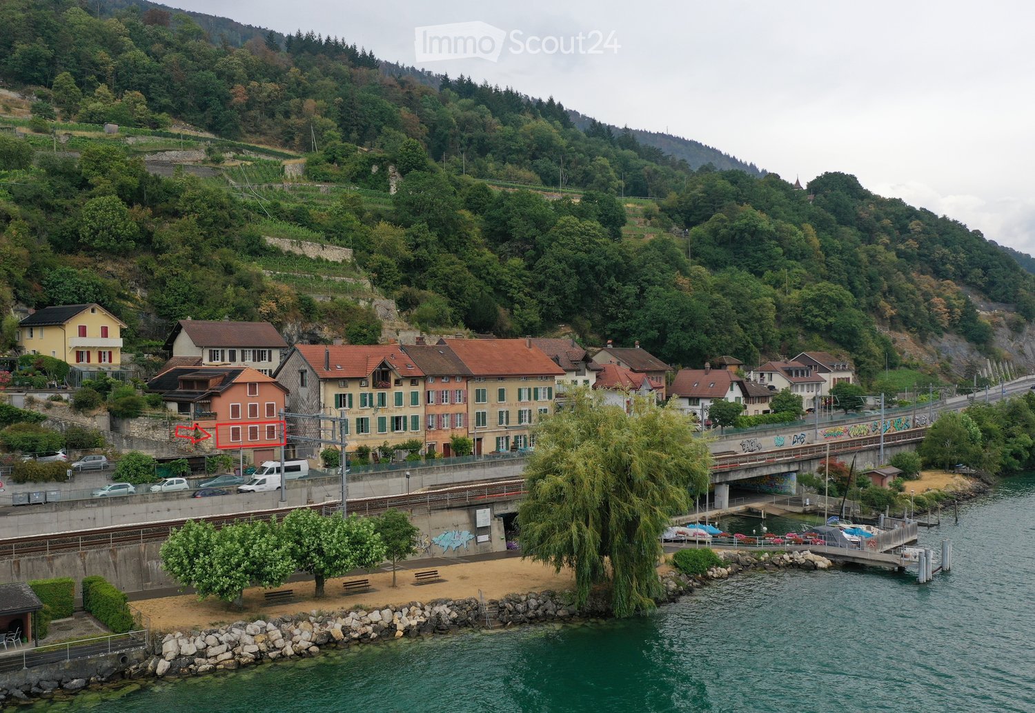 A cluster of residential buildings with varied architectural styles, situated along a riverbank with a bridge and a small dock area visible.