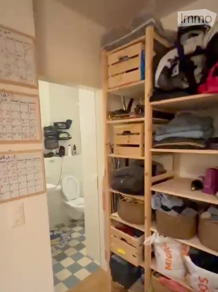 A small bathroom with a toilet, checkered floor, and a wooden shelf unit filled with storage boxes and folded clothes. Calendars are visible on the wall.
