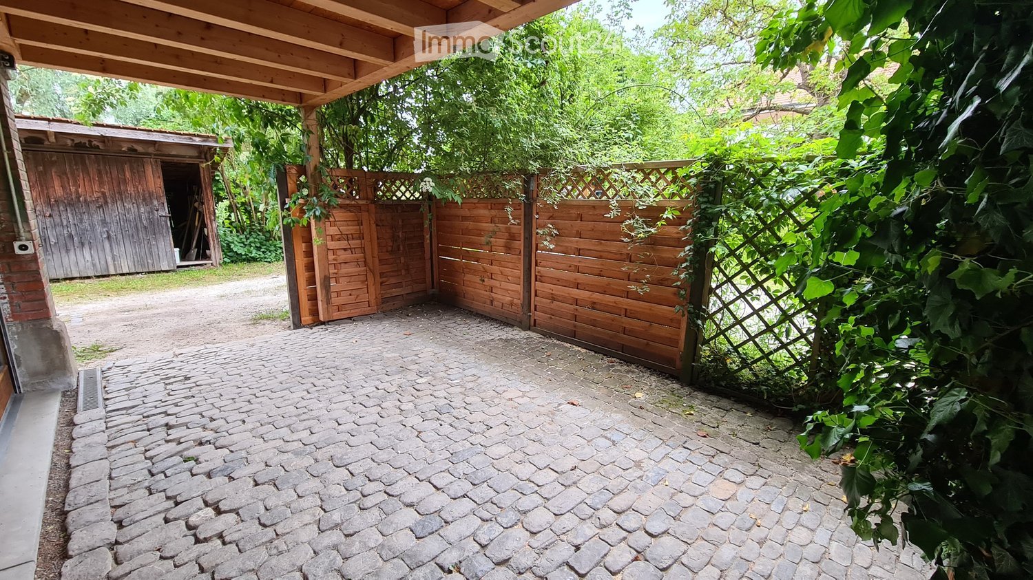 A cobblestone pathway leads to a wooden gate, with a wooden shed visible in the background and lush greenery on either side.
