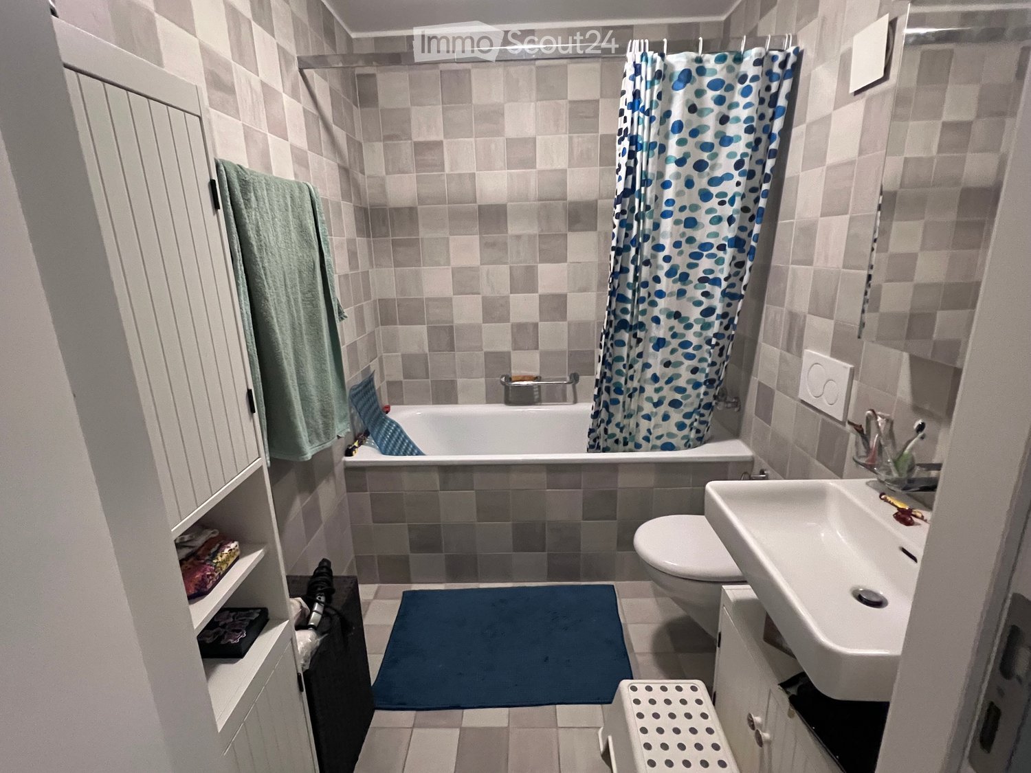 Modern bathroom with a bathtub, toilet, sink, and a small cabinet. Features a blue and white patterned shower curtain, a green towel, and a blue bathroom rug.