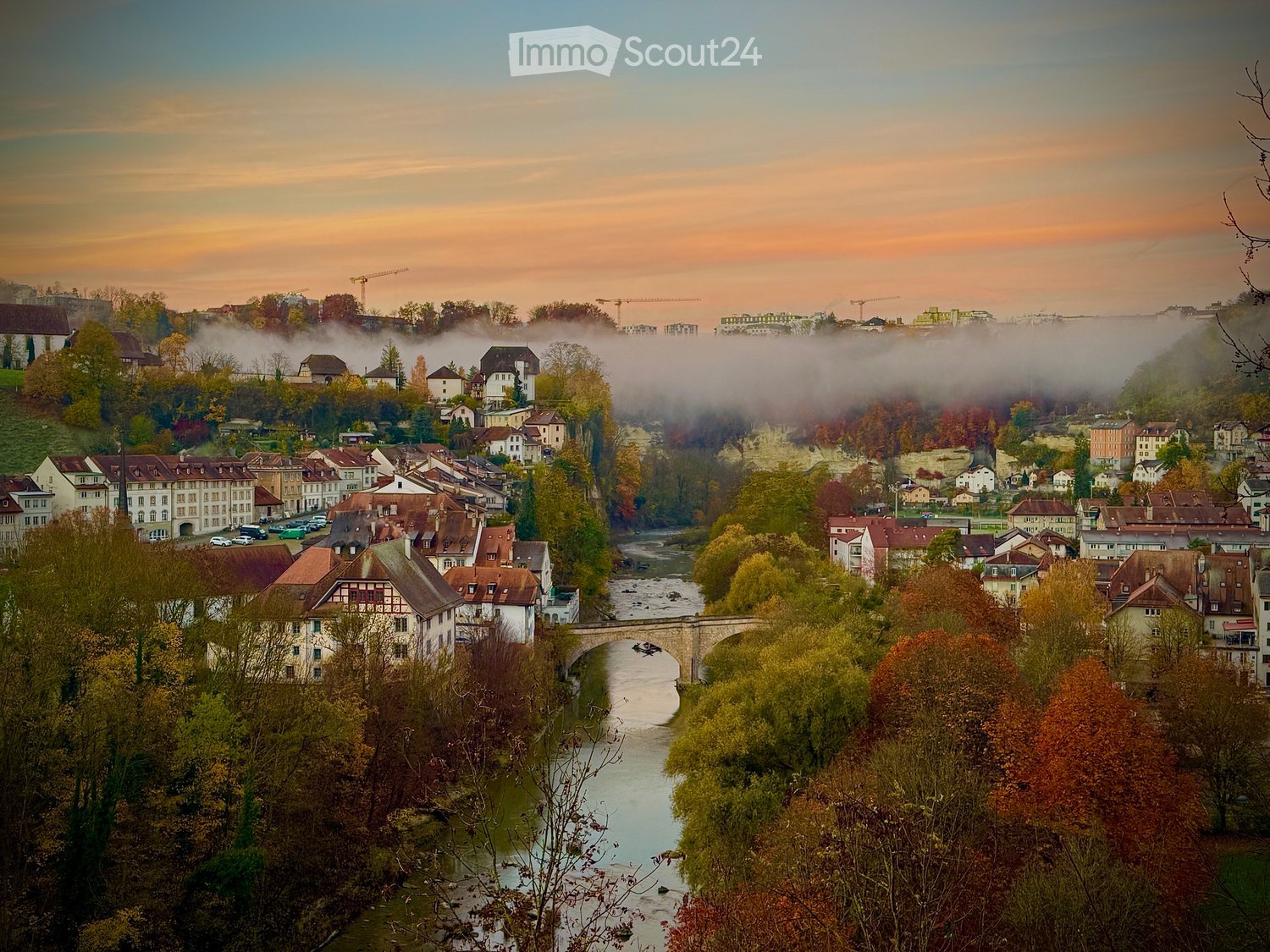 A panoramic view of a town with a river running through it, surrounded by houses and trees, with a bridge crossing the river and cranes visible in the distance.
