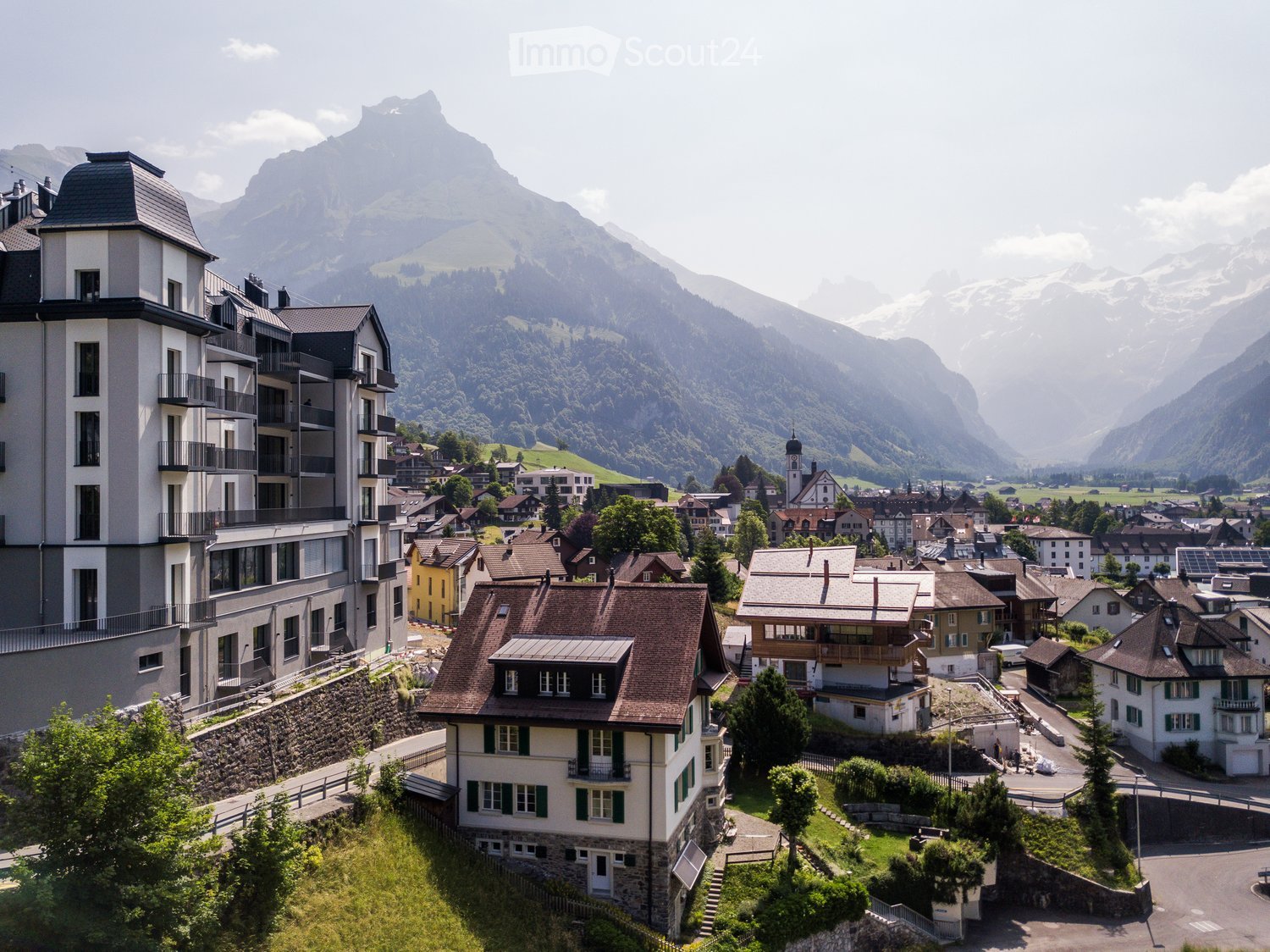 Aerial view of a village nestled in a valley with snow-capped mountains in the background. The village features a mix of residential and commercial buildings, including multi-story apartment complexes, traditional houses with pitched roofs, and a church t