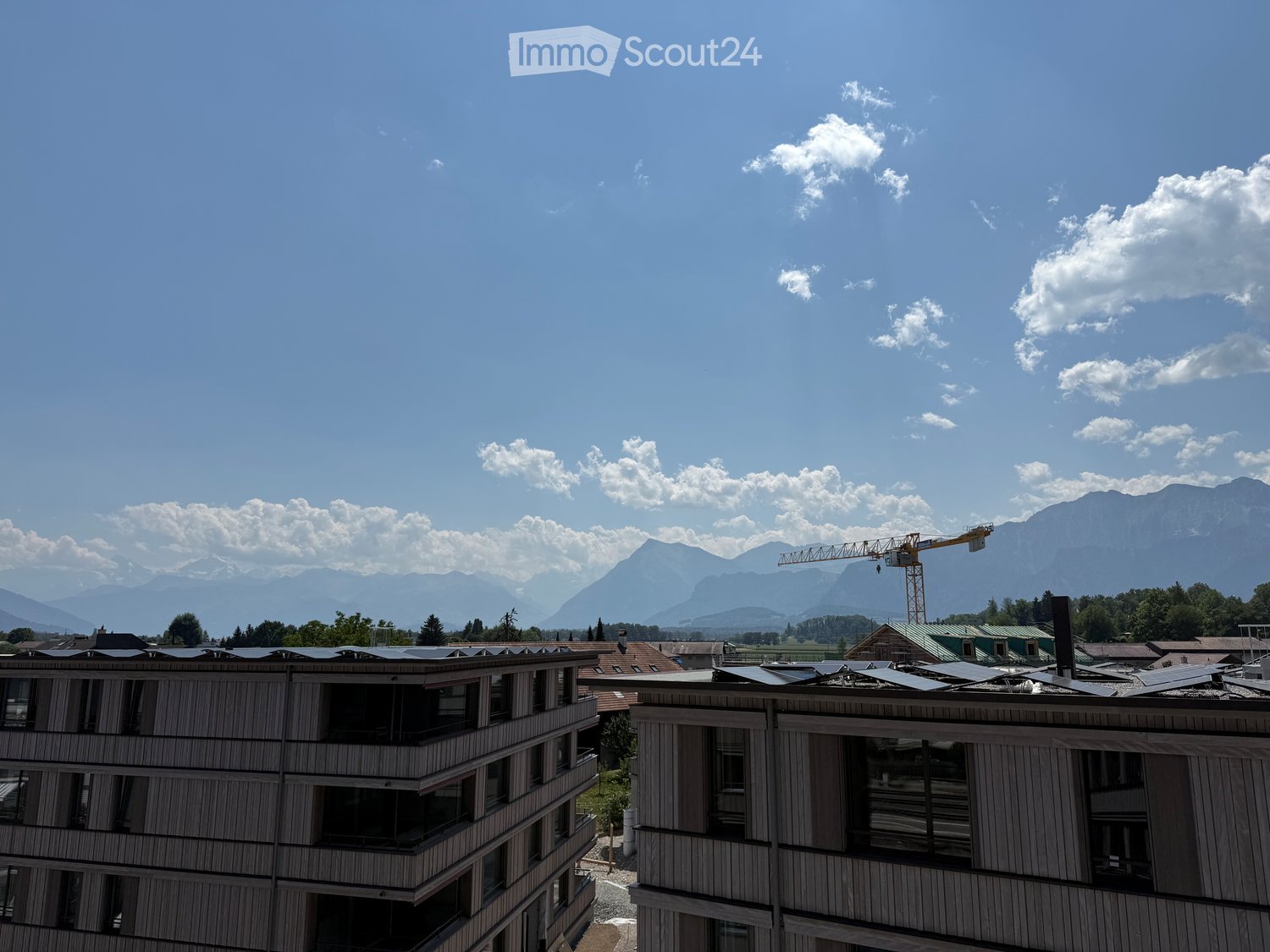 Modern multi-story building with large windows and solar panels on the roof, surrounded by greenery and mountains under a partly cloudy sky.