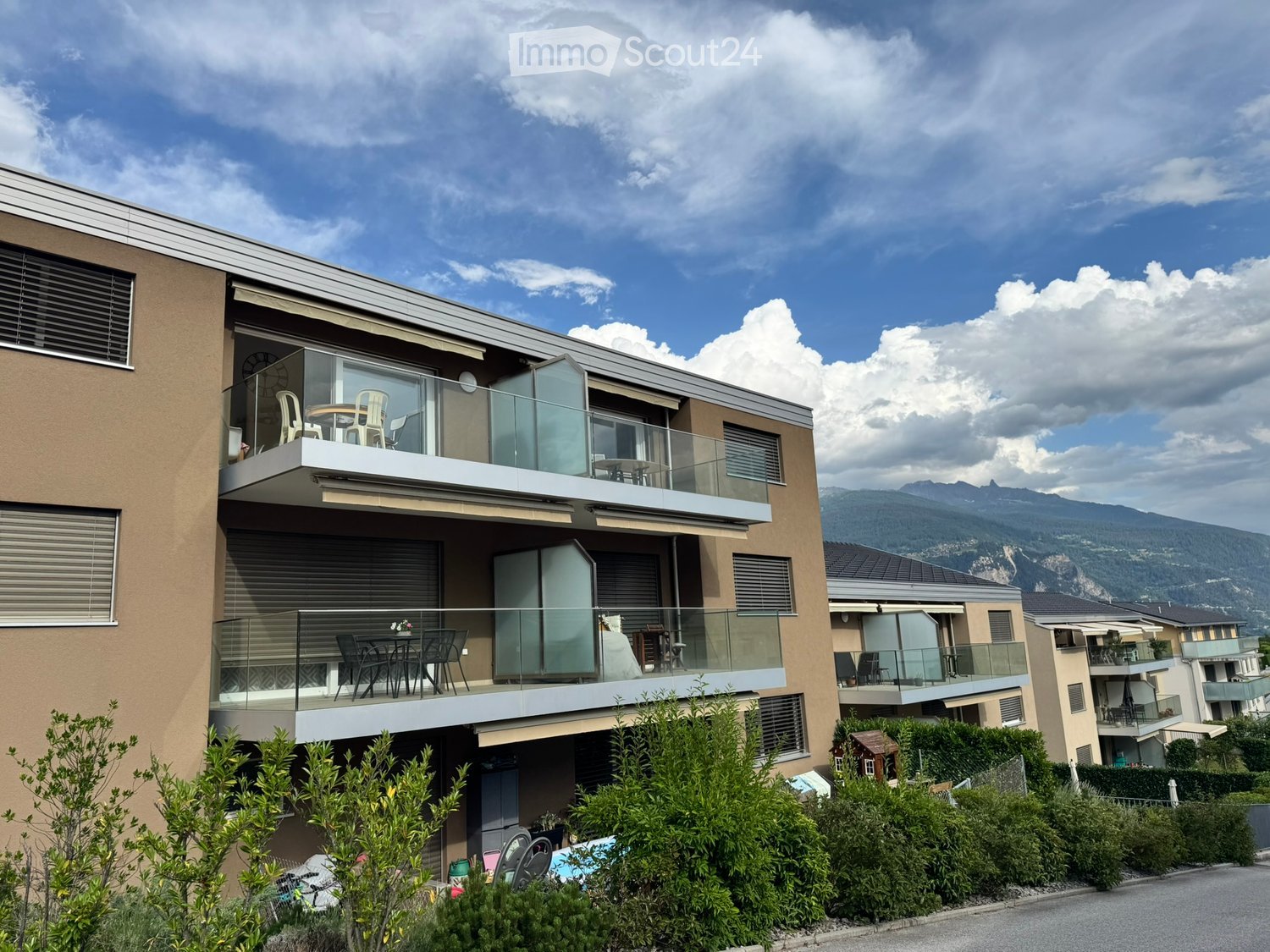 Modern two-story building with balconies, glass railings, and outdoor furniture. Features a swimming pool in the foreground and scenic mountain views in the background.