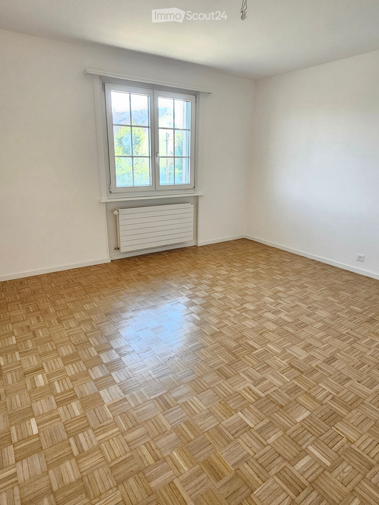 Empty room with wooden herringbone flooring, white walls, large window with white frame, and radiator below the window.