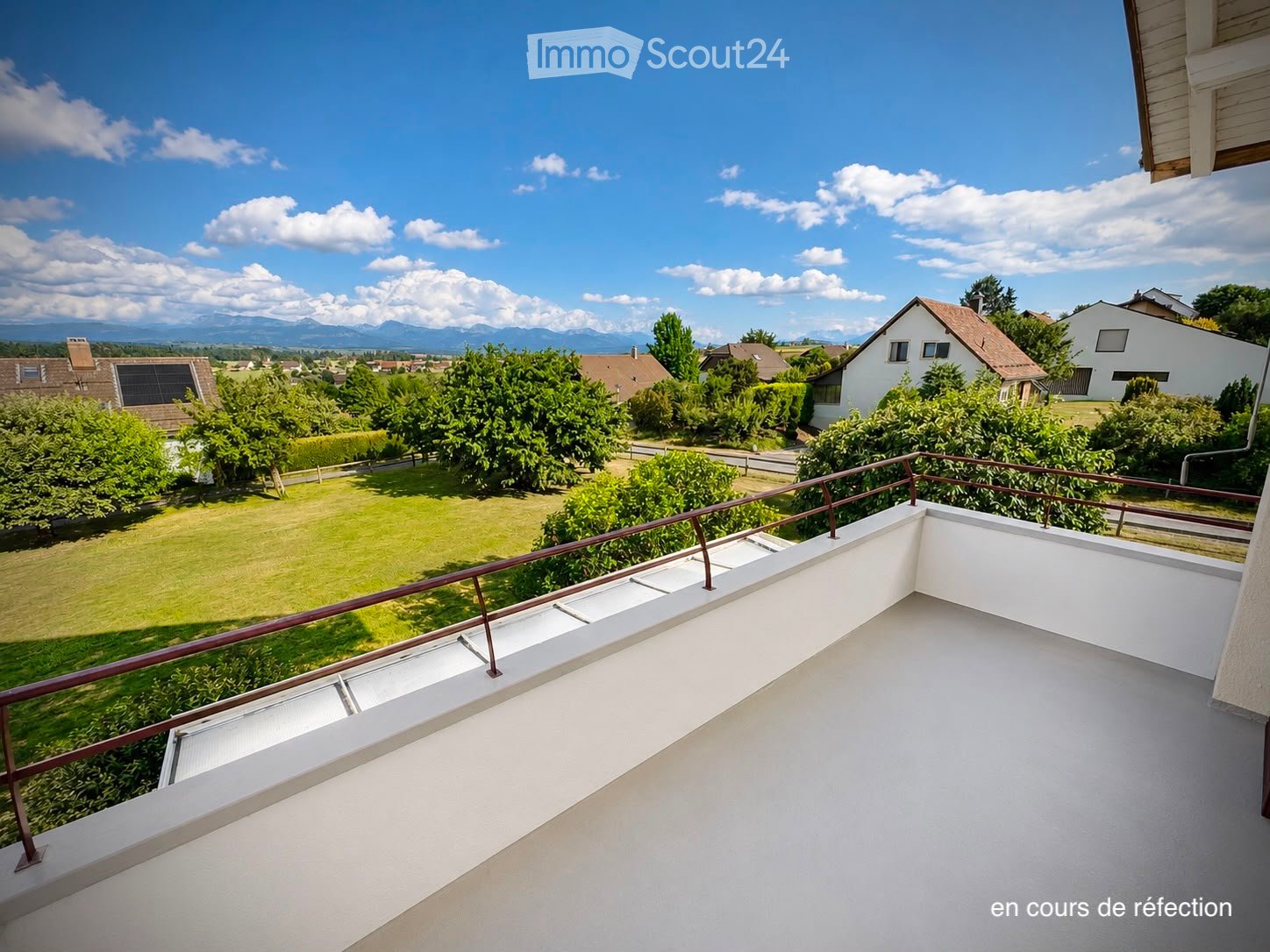 A modern balcony with a white floor and metal railing, overlooking a green lawn, trees, and distant mountains under a clear blue sky.