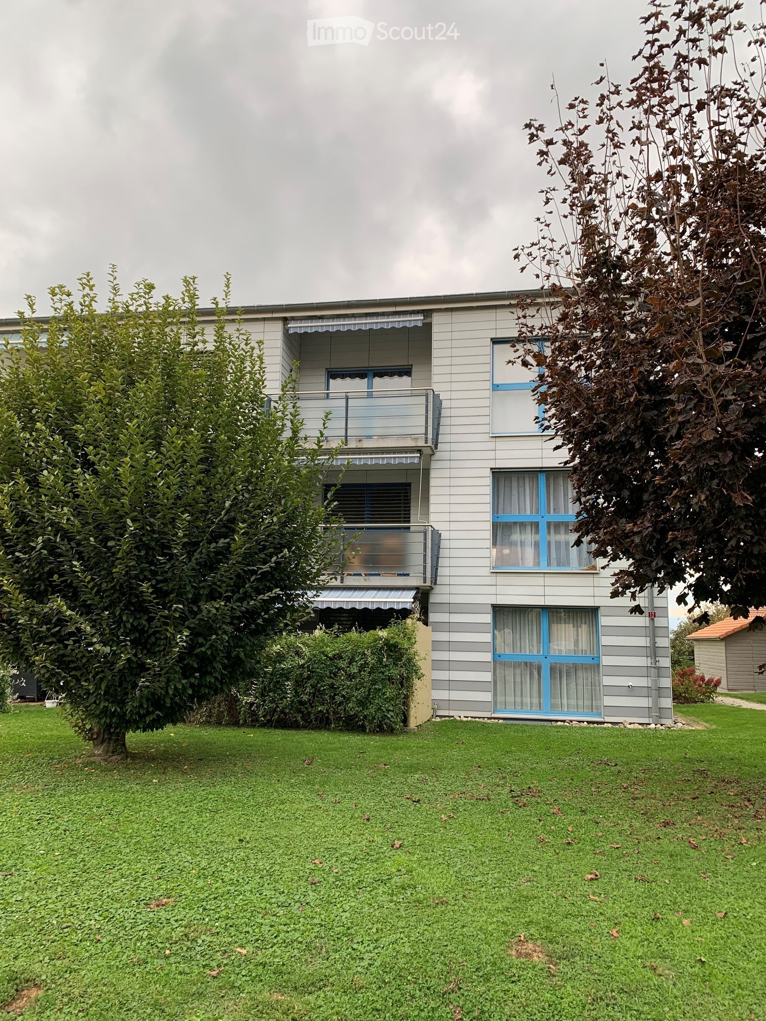 Two-story house with grey siding, blue-framed windows, and balconies. The house is surrounded by a well-maintained lawn and trees.
