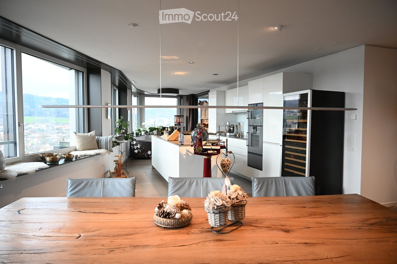 Open-plan living and dining area with a modern kitchen, large windows offering a scenic view, a wooden dining table with chairs, a sofa, and decorative items.