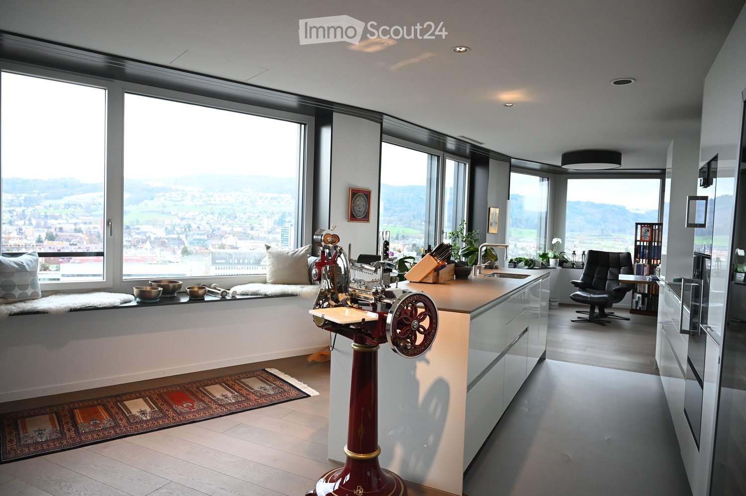 Modern kitchen with large windows offering a scenic view, featuring a kitchen island, cabinets, a sink, a stool, a chair, and a framed picture on the wall.