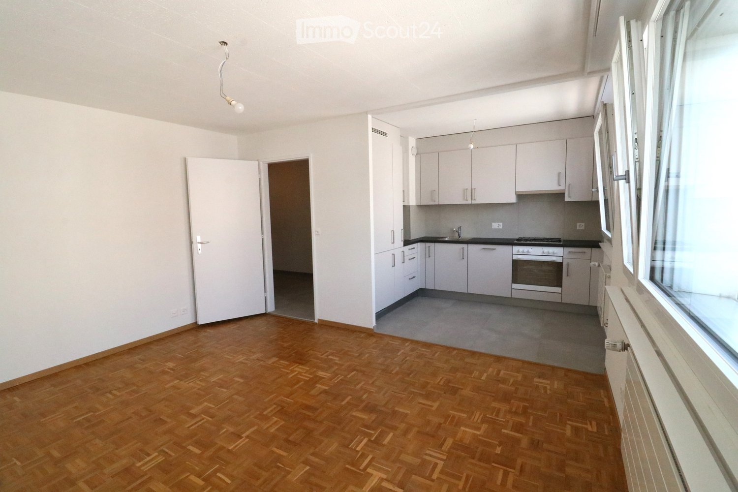Empty room with wooden flooring, white walls, and a doorway leading to a kitchen area with white cabinets and appliances.