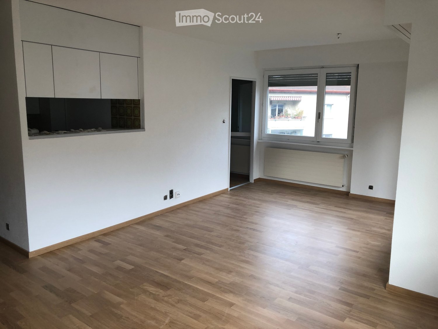 Empty room with light wooden flooring, white walls, and a large window with blinds. The room features built-in cabinets and a radiator under the window.