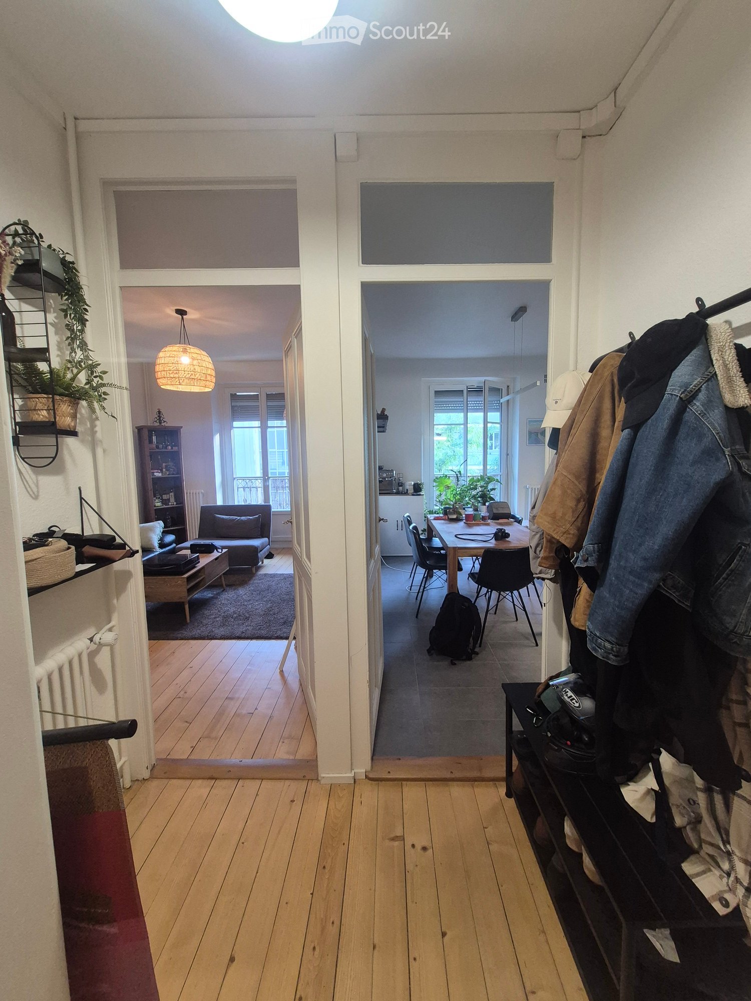 A well-lit hallway with wooden flooring, leading to a living room and a dining area. The living room features a sofa, coffee table, and a hanging lamp. The dining area includes a table, chairs, and a backpack on the floor.