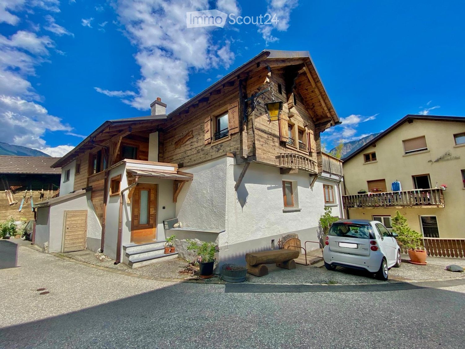 A two-story wooden house with a balcony, surrounded by other houses and a small car parked in front.