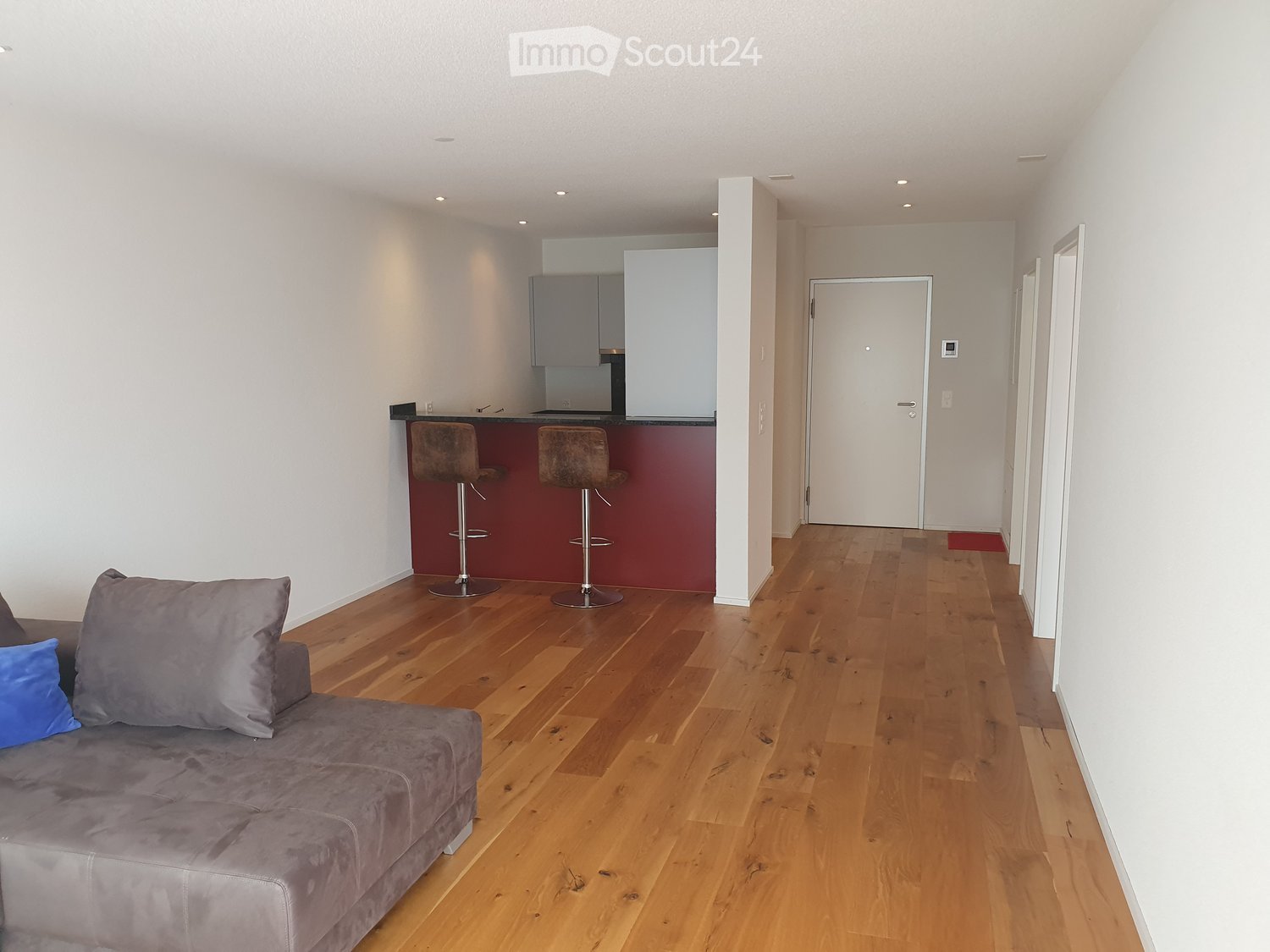 Modern living room with a grey sofa, two bar stools, and a kitchen area with cabinets.