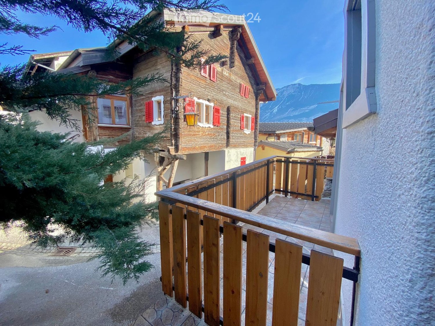 Balcony with wooden railing, overlooking a stone house with red shutters and a view of mountains.