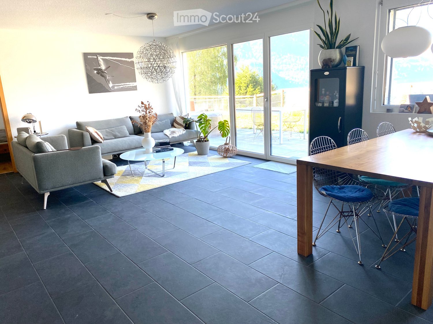 Modern living and dining area with grey sofa, coffee table, dining table with chairs, large windows, and decorative items.