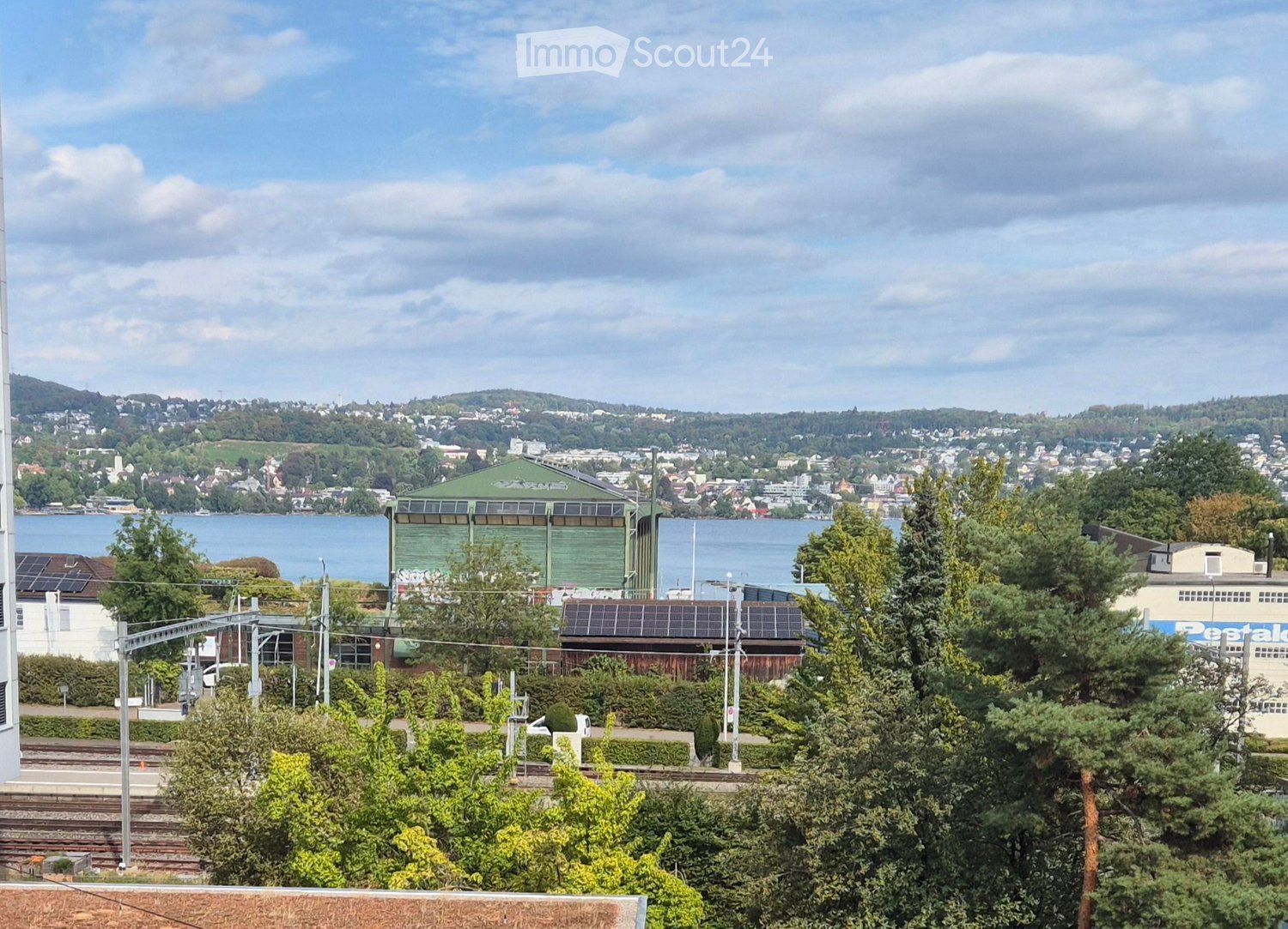 A panoramic view of a cityscape with a body of water in the background, featuring a mix of residential and industrial buildings, trees, and a clear sky.