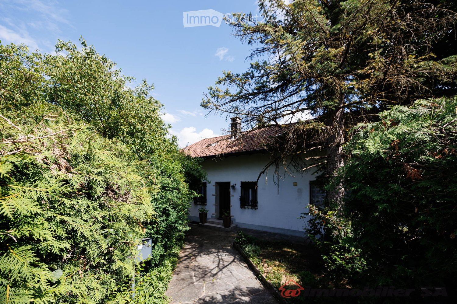 A small, single-story white house with a tiled roof, surrounded by lush greenery and trees. The house features a front door with a small porch and windows, and is accessed via a stone pathway.