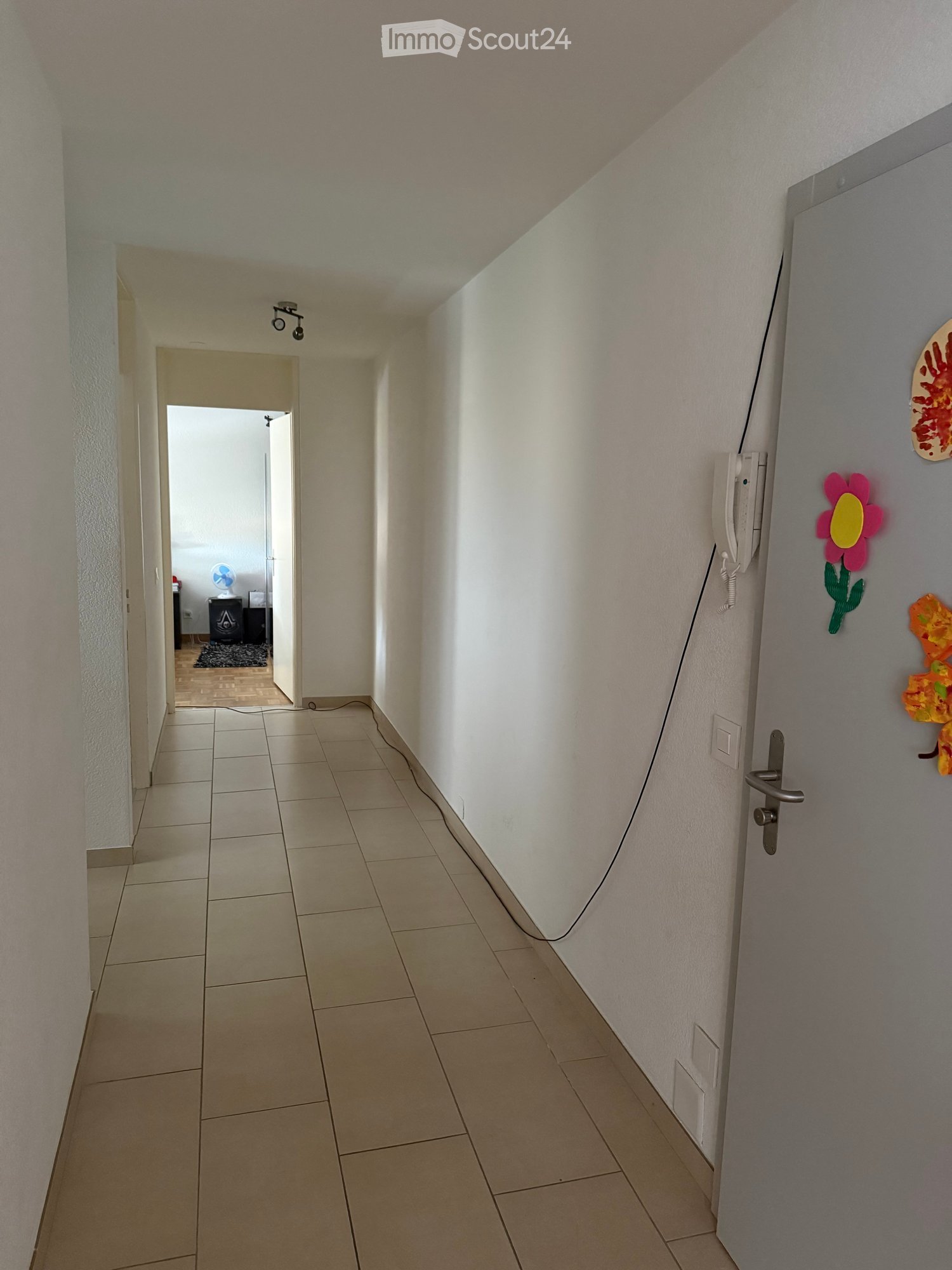 A clean, tiled hallway with white walls, a closed door on the right, and an open doorway leading to another room on the left. The hallway has a light fixture on the ceiling and a small fan visible in the adjacent room.