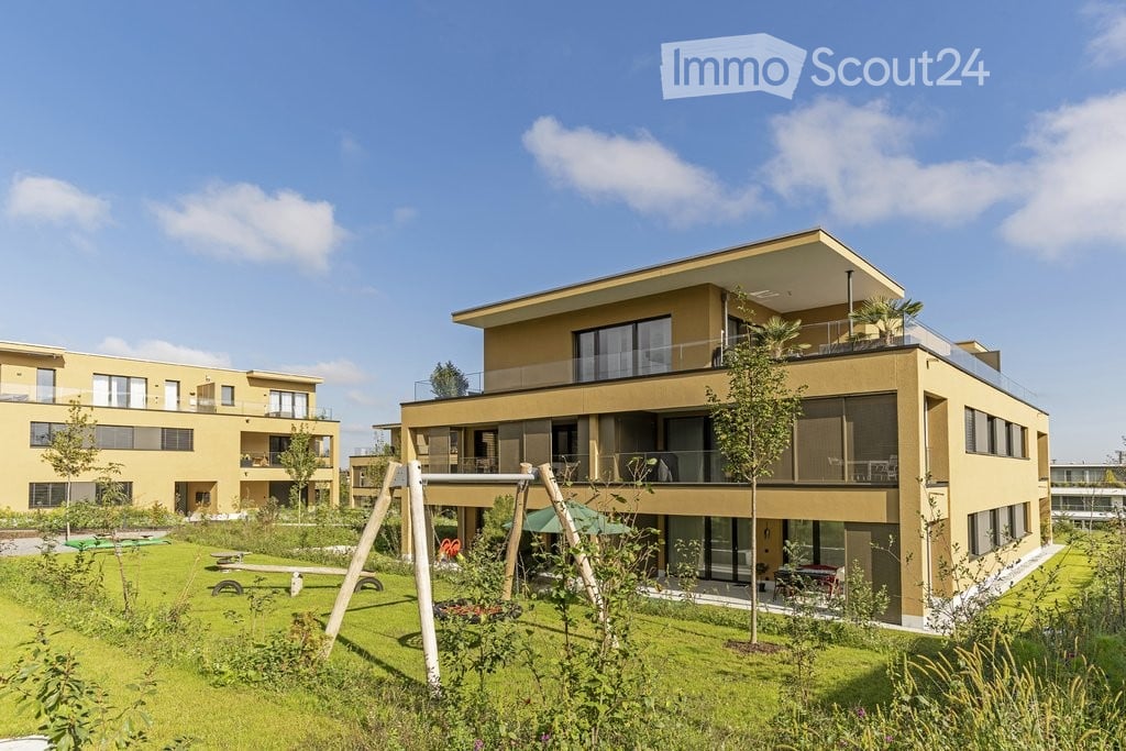 Modern two-story residential building with large windows, balconies, and a landscaped garden area with playground equipment.