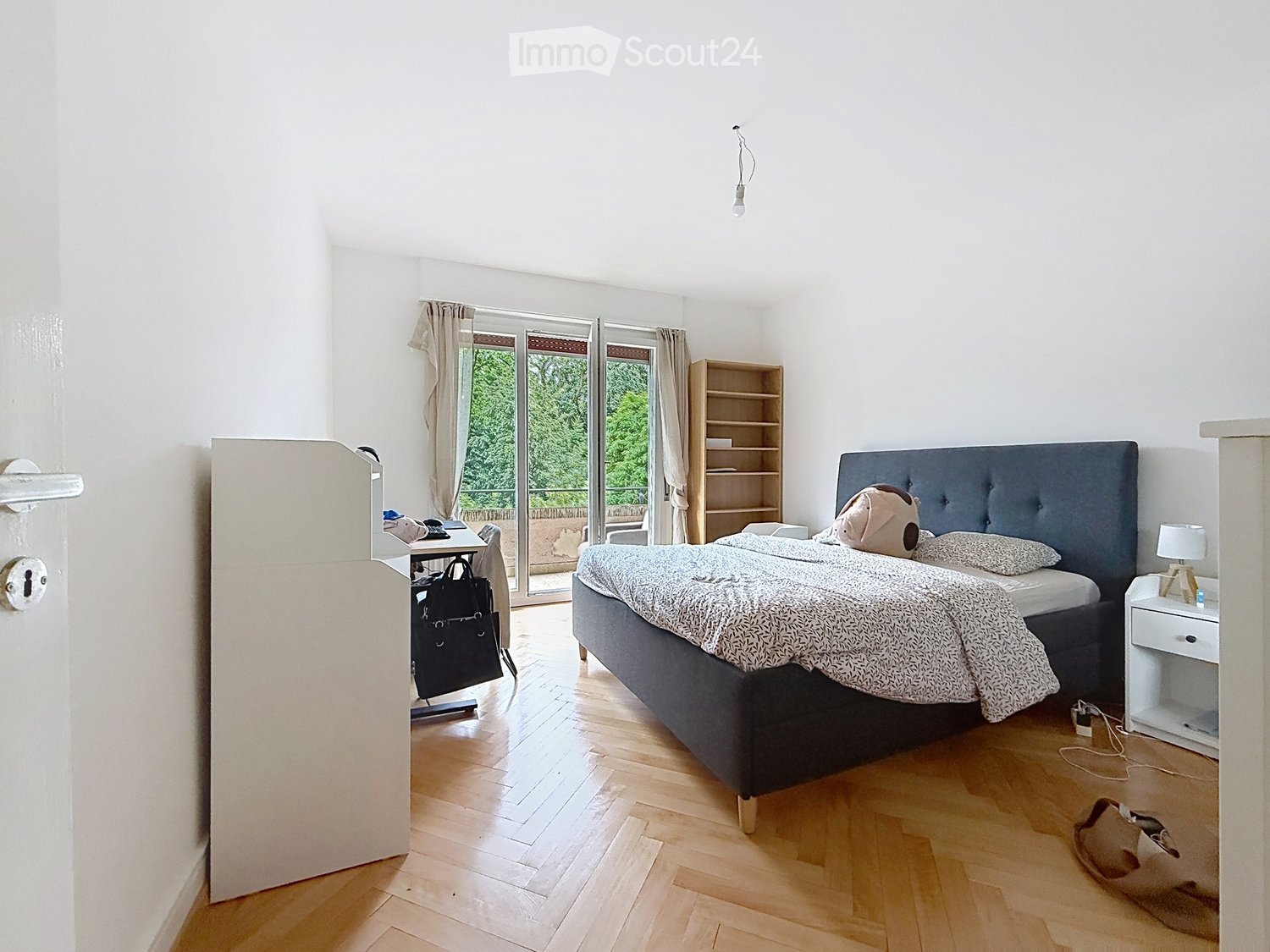 Bright bedroom with wooden flooring, large bed with patterned bedding, nightstand with lamp, bookshelf, and a chair with a handbag nearby. Large windows with curtains lead to a balcony.