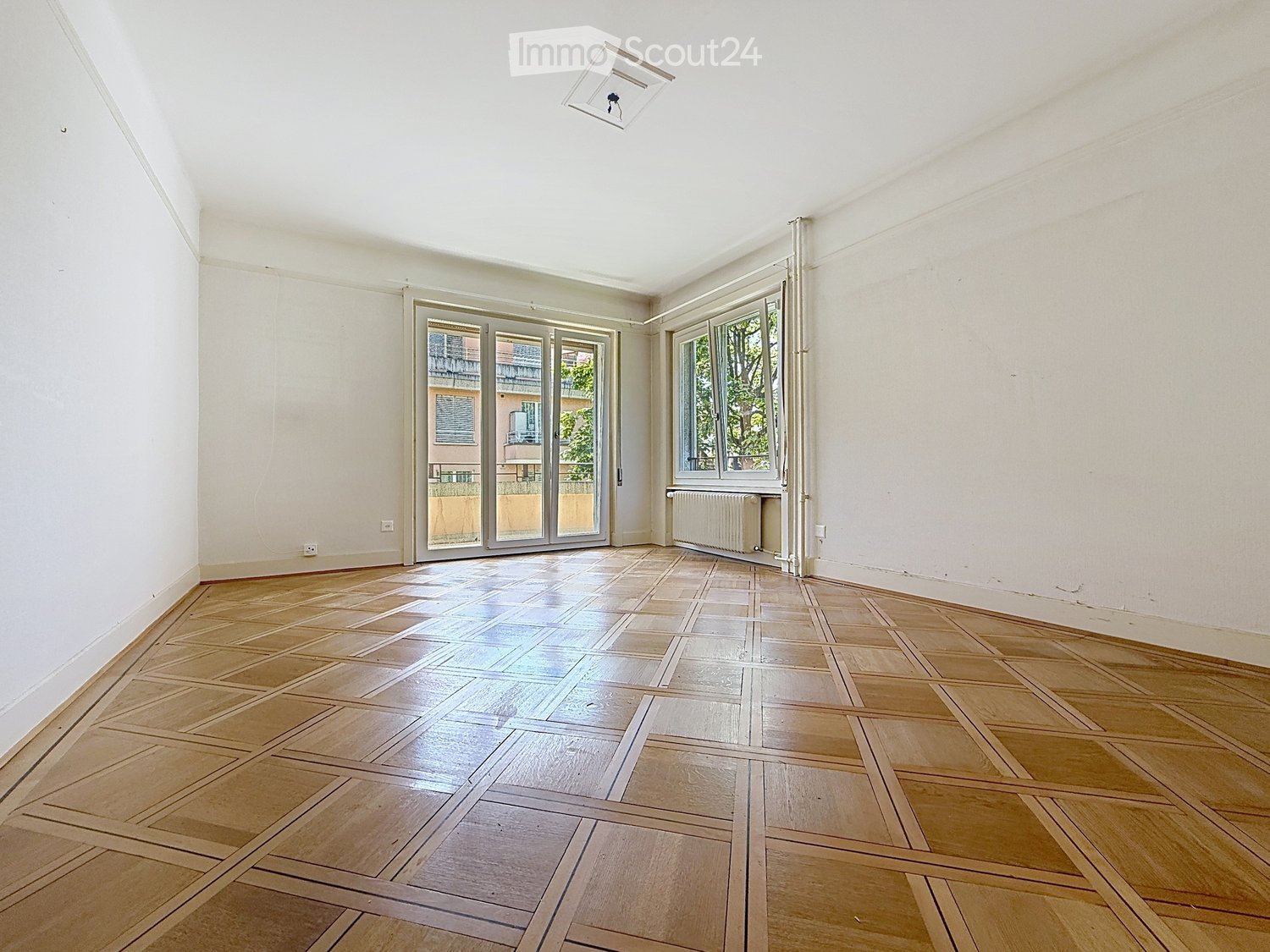 Empty room with wooden parquet flooring, white walls, large windows, and a sliding glass door leading to a balcony.
