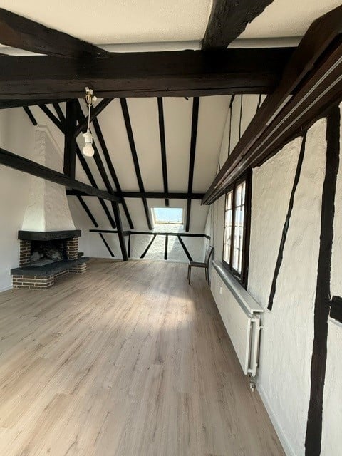 An attic space with exposed wooden beams, a fireplace, and wooden flooring. The room is empty, with a single window and a small skylight providing natural light.