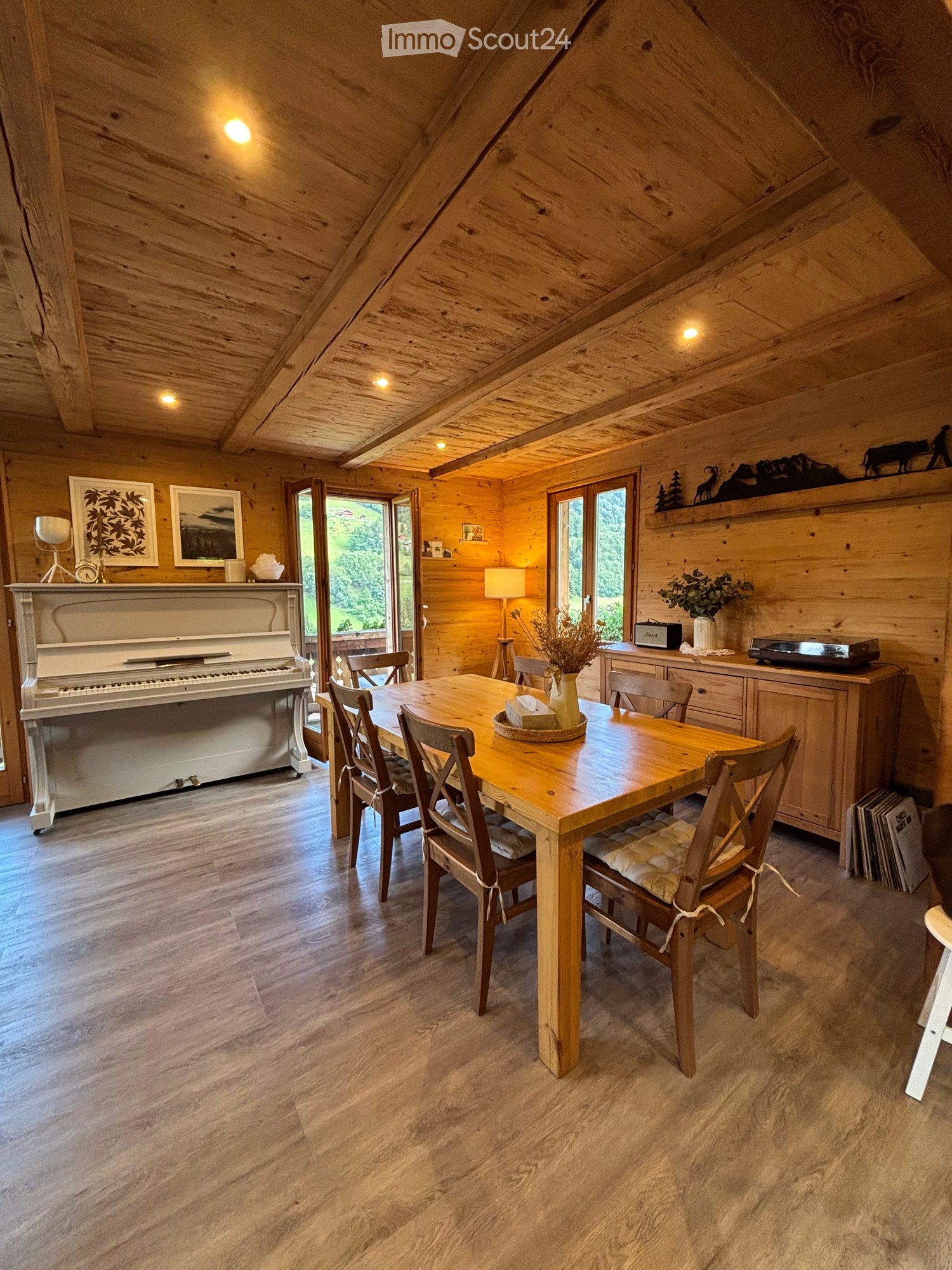 Cozy wooden-paneled room with a dining table, chairs, piano, and decorative items. Large windows offer a scenic view.