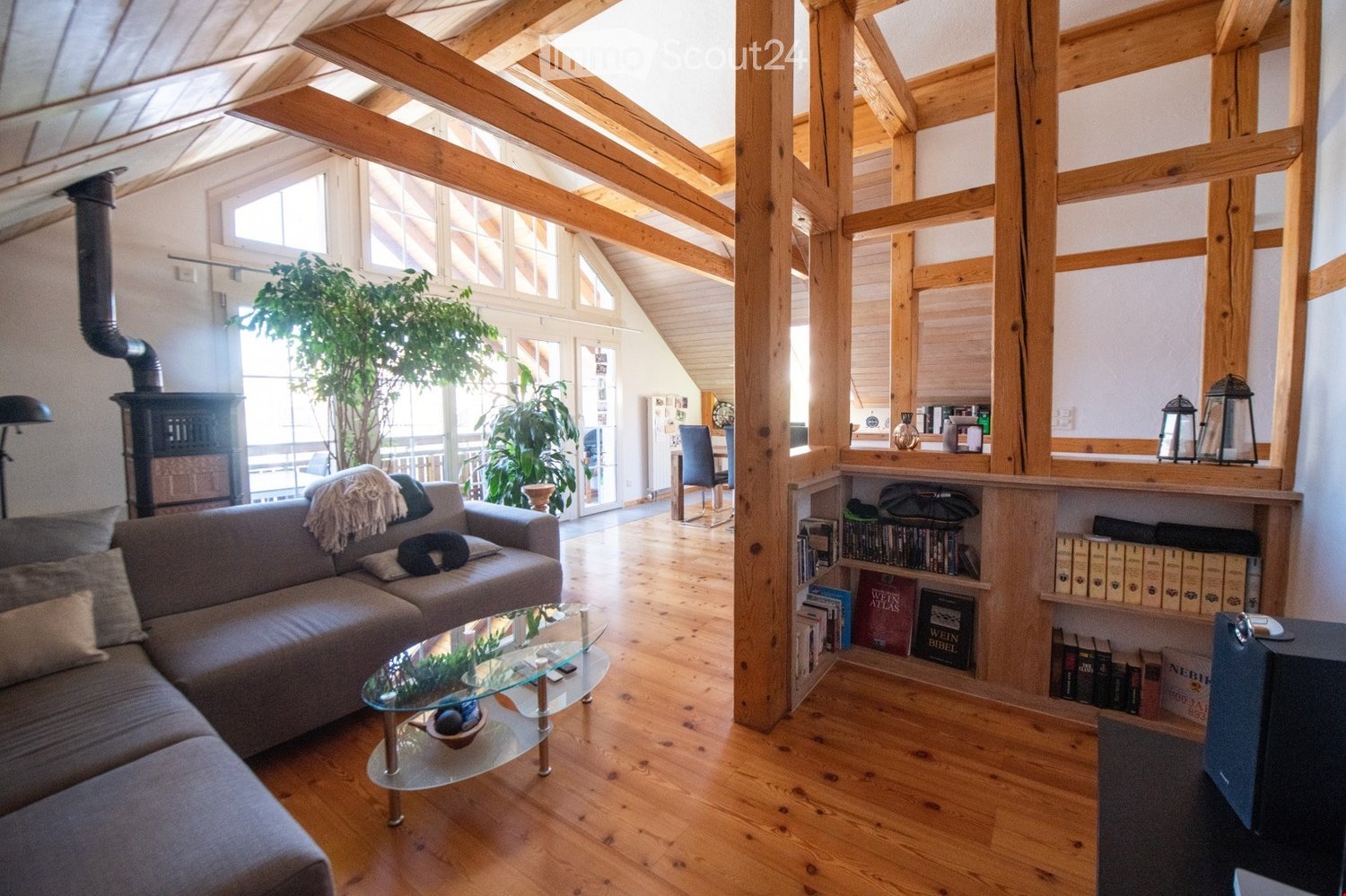 Bright living room with wooden beams, large windows, and skylights. Features a grey sofa, glass coffee table, bookshelf, dining area with a table and chairs, and a fireplace.