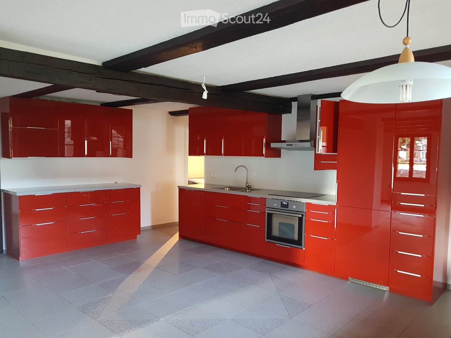 Modern kitchen with red cabinets, stainless steel oven, sink, and faucet, featuring exposed wooden beams and a pendant light.