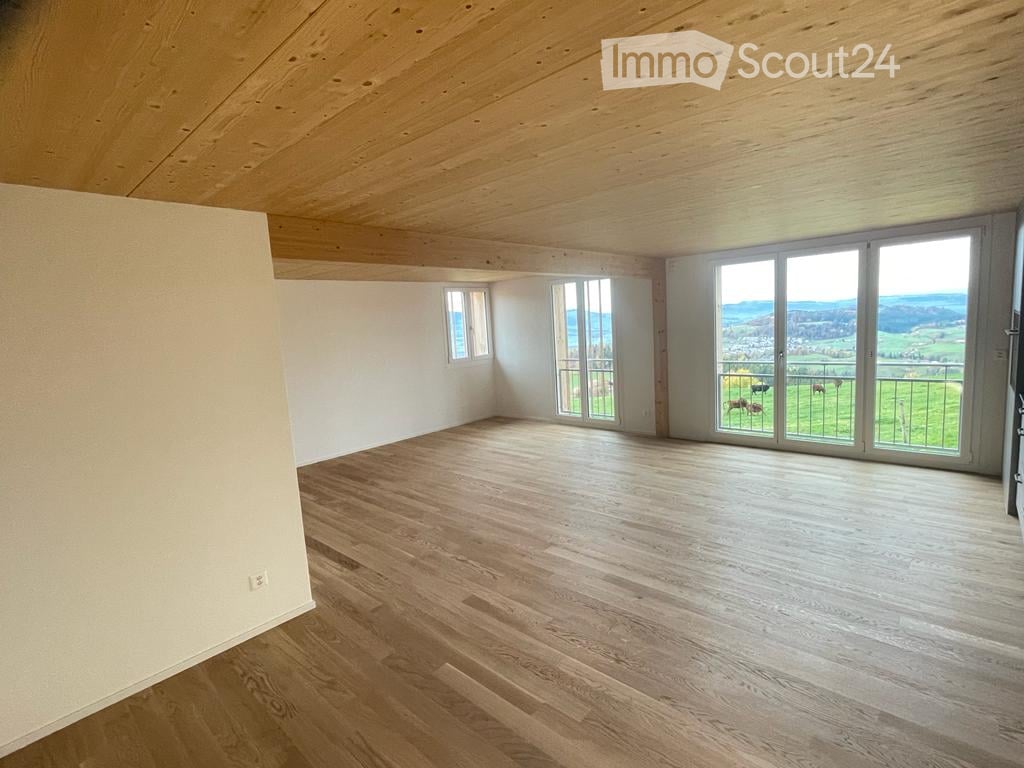 Empty room with wooden floor, white walls, large windows, and a view of a green landscape.