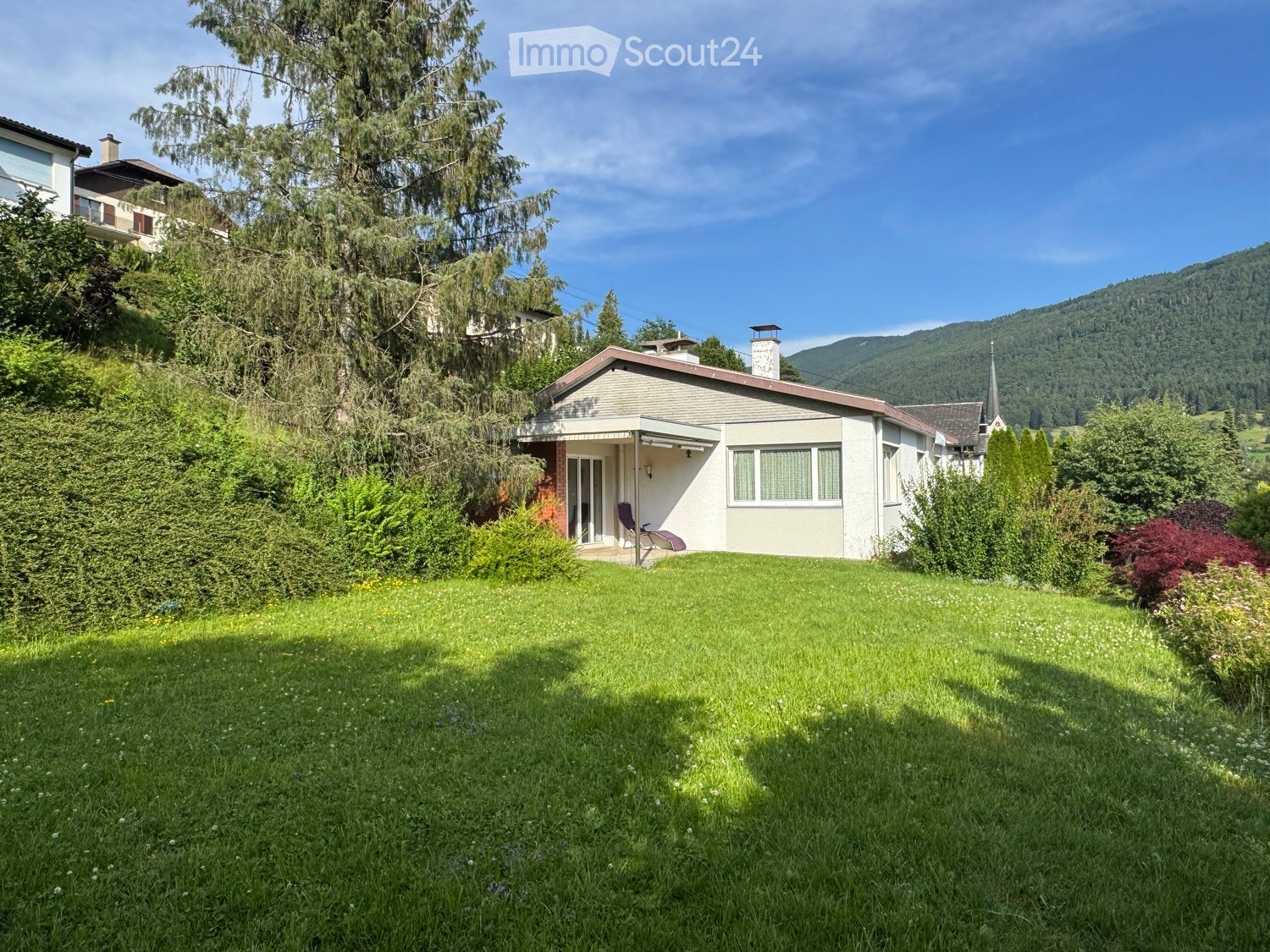 Single-story house with a sloped roof, surrounded by a well-maintained lawn and trees, with a scenic mountain view in the background.