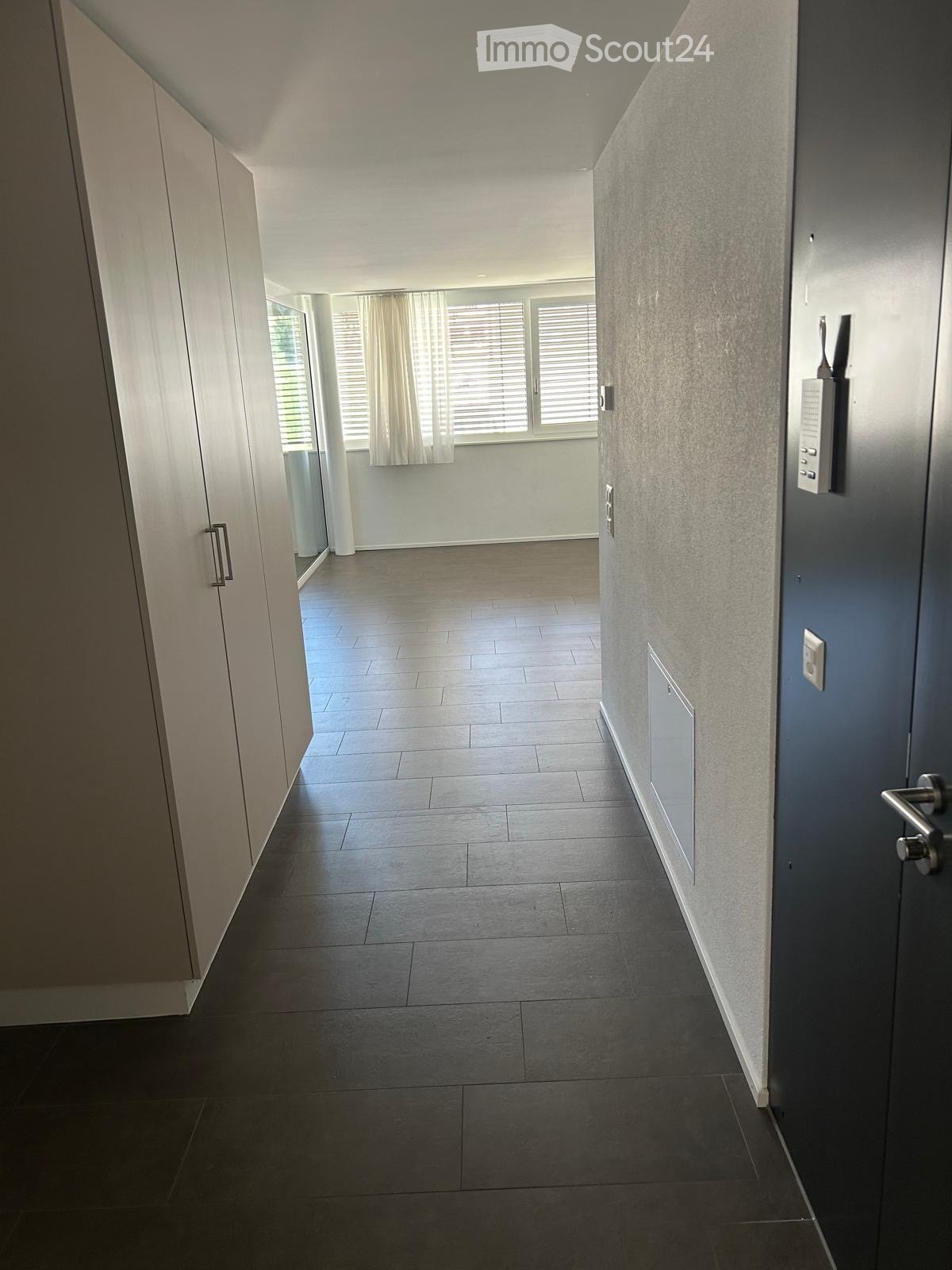 Modern hallway with dark tiled flooring, white walls, and a door leading to a room with large windows and light-colored curtains.