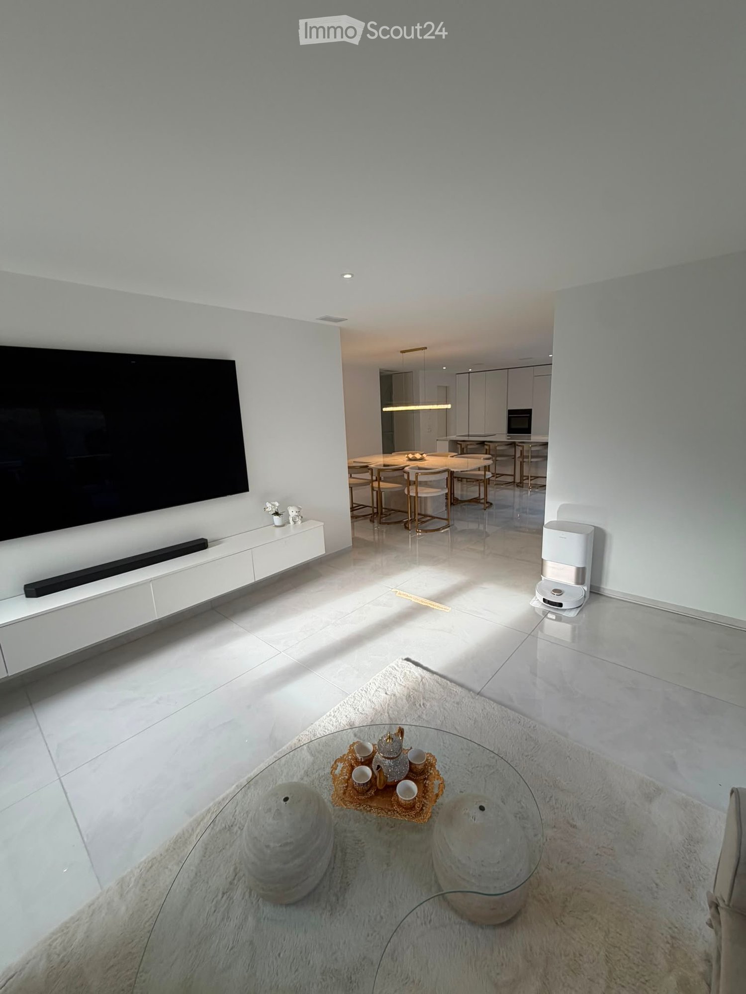 Modern living room with white walls, large TV, glass coffee table, decorative tea set, and a dining area with chairs and a table in the background.