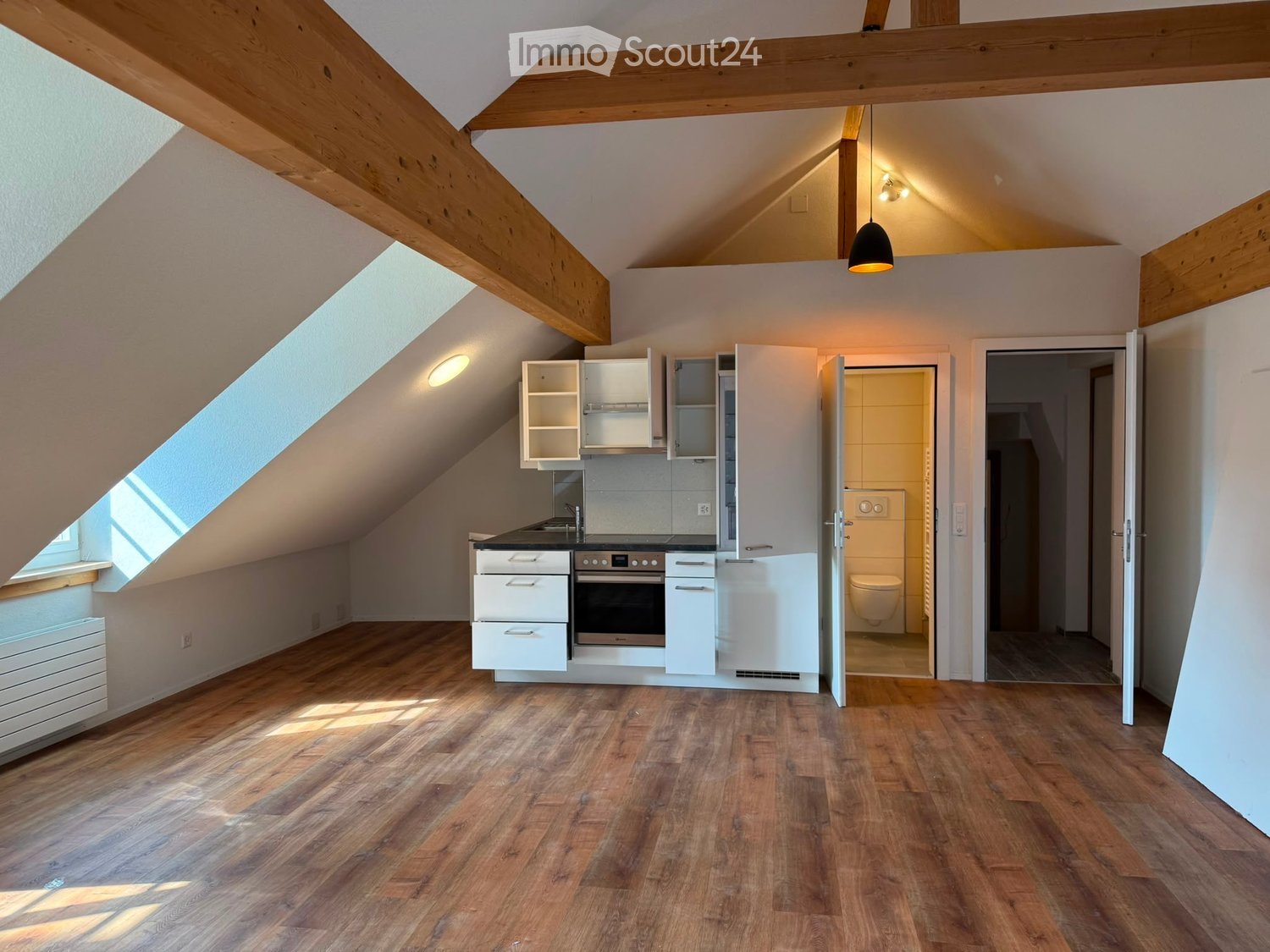 Bright attic space with wooden floors, white cabinets, an oven, and a small bathroom with a toilet.