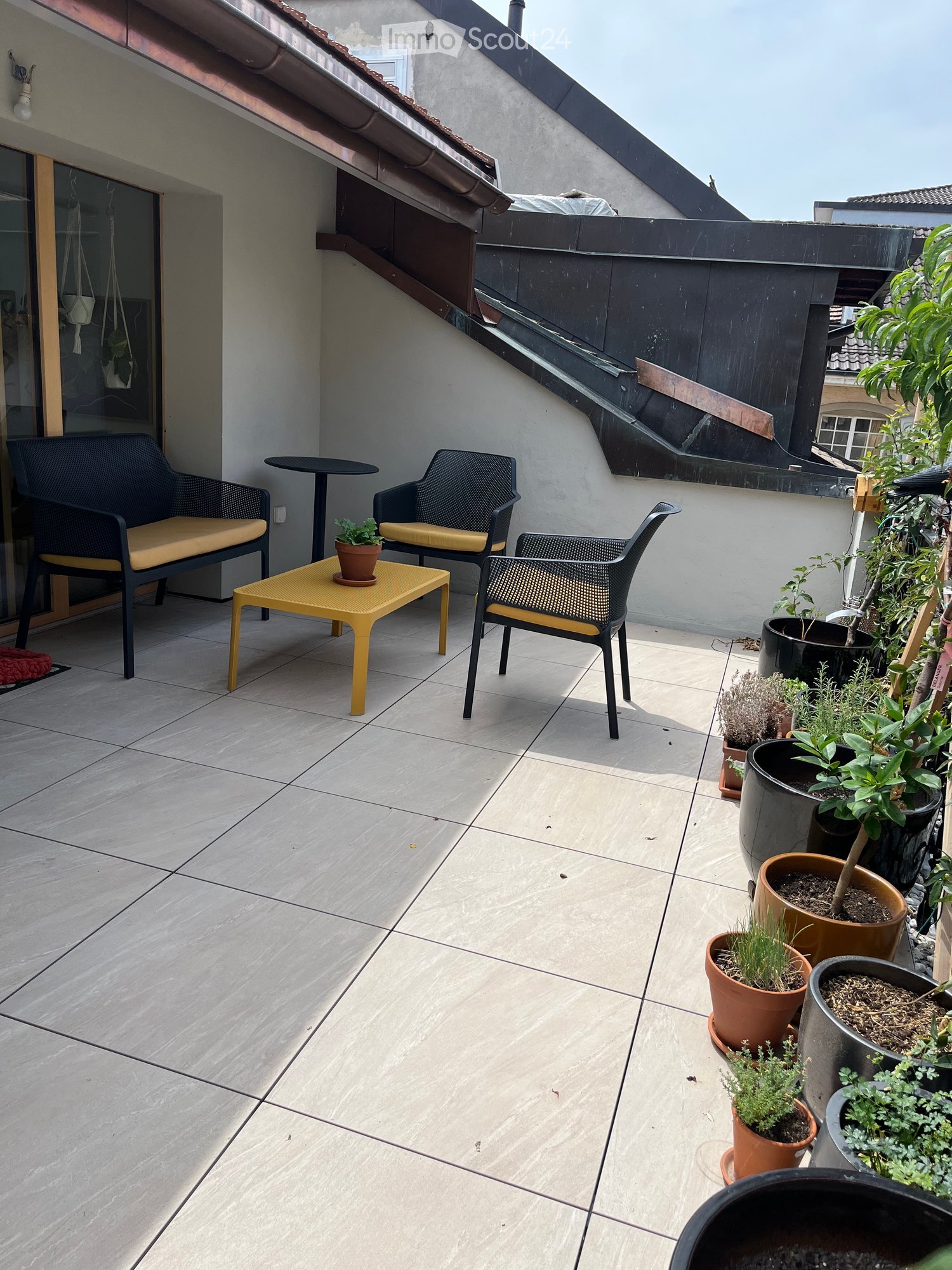 Balcony with tiled floor, several potted plants, and outdoor furniture including chairs and a small table.