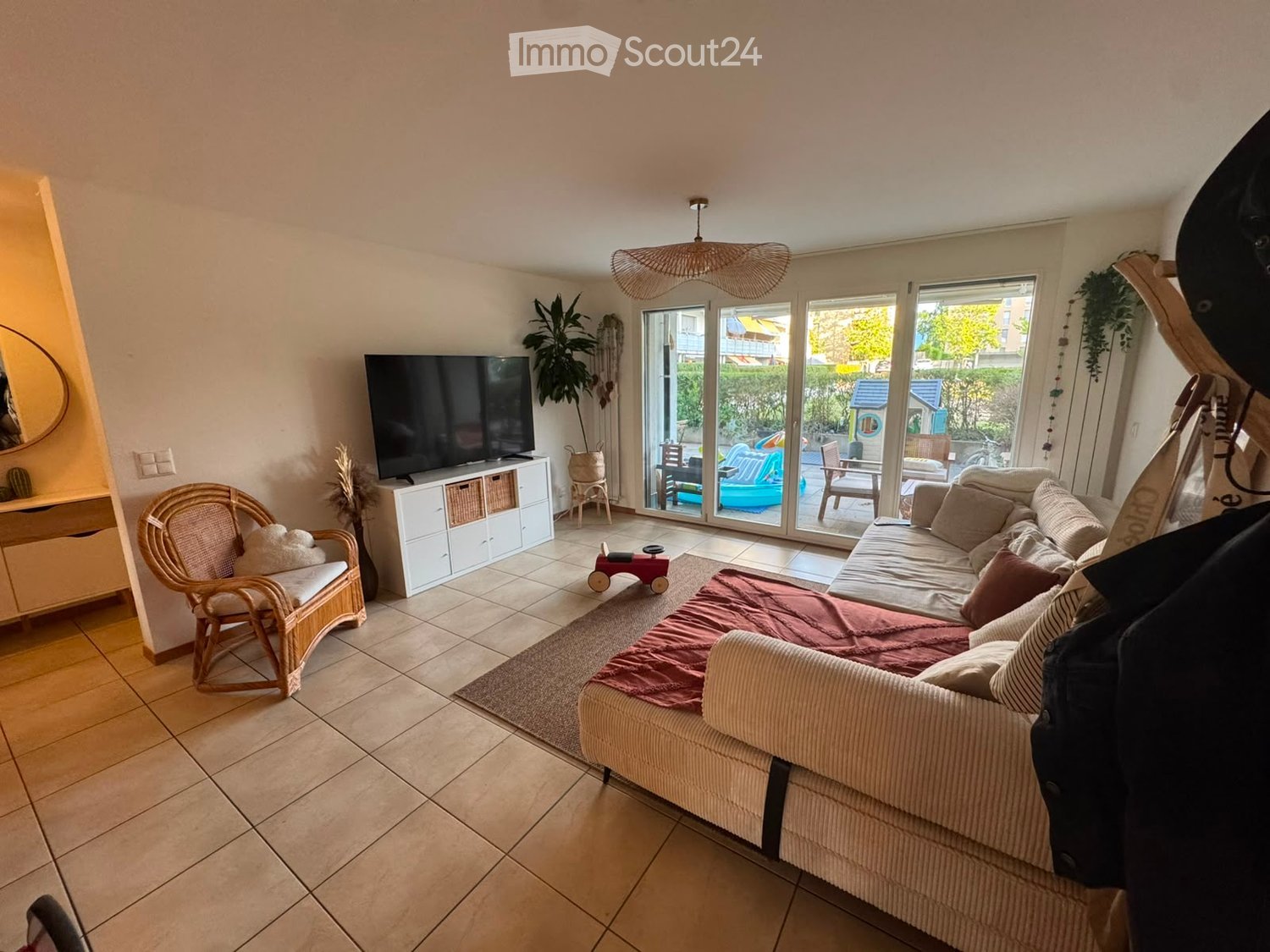 Bright living room with large sliding glass doors leading to a patio, a beige sofa with throw pillows, a wicker hanging lamp, a wicker chair, a TV on a white stand, and a potted plant near the entrance.