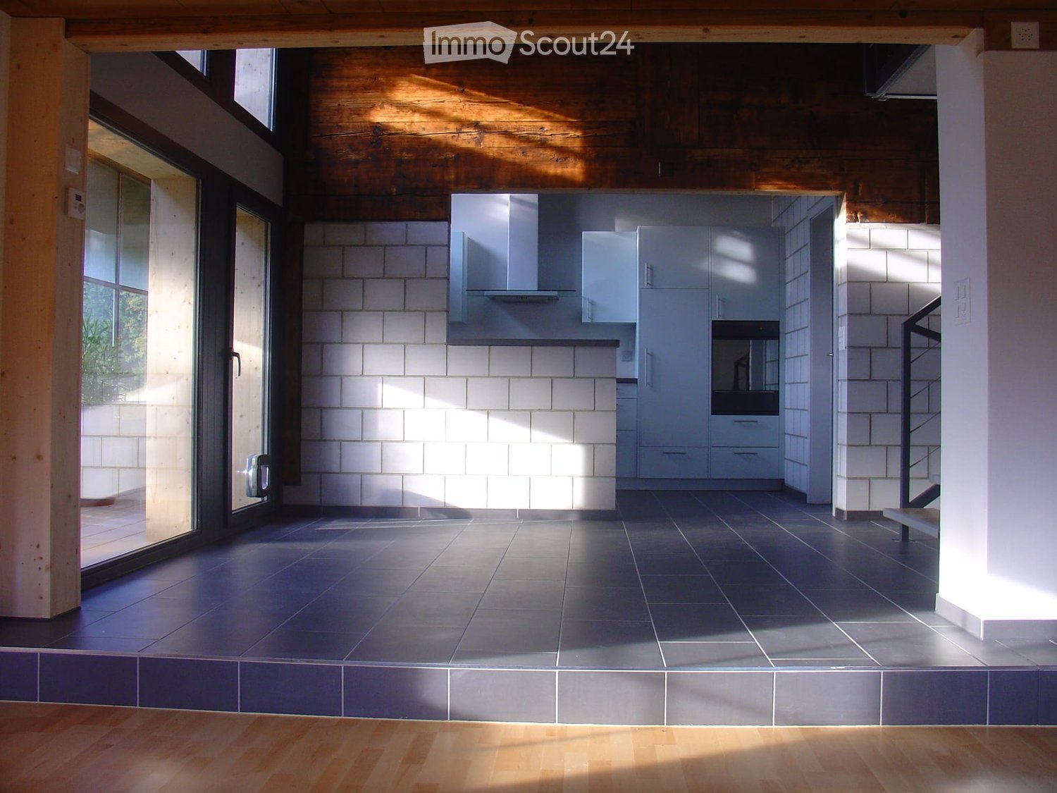 Modern kitchen with white tiled walls, dark tiled floor, white cabinets, and an oven. Large windows and wooden beams on the ceiling.